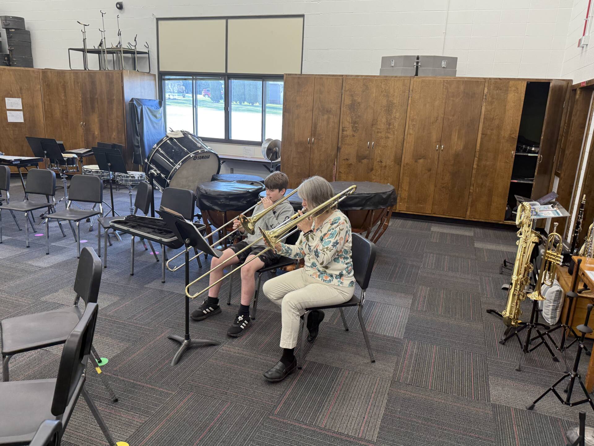 A student and a teacher sit in a music room playing trombones, surrounded by chairs, drums, and instrument stands.
