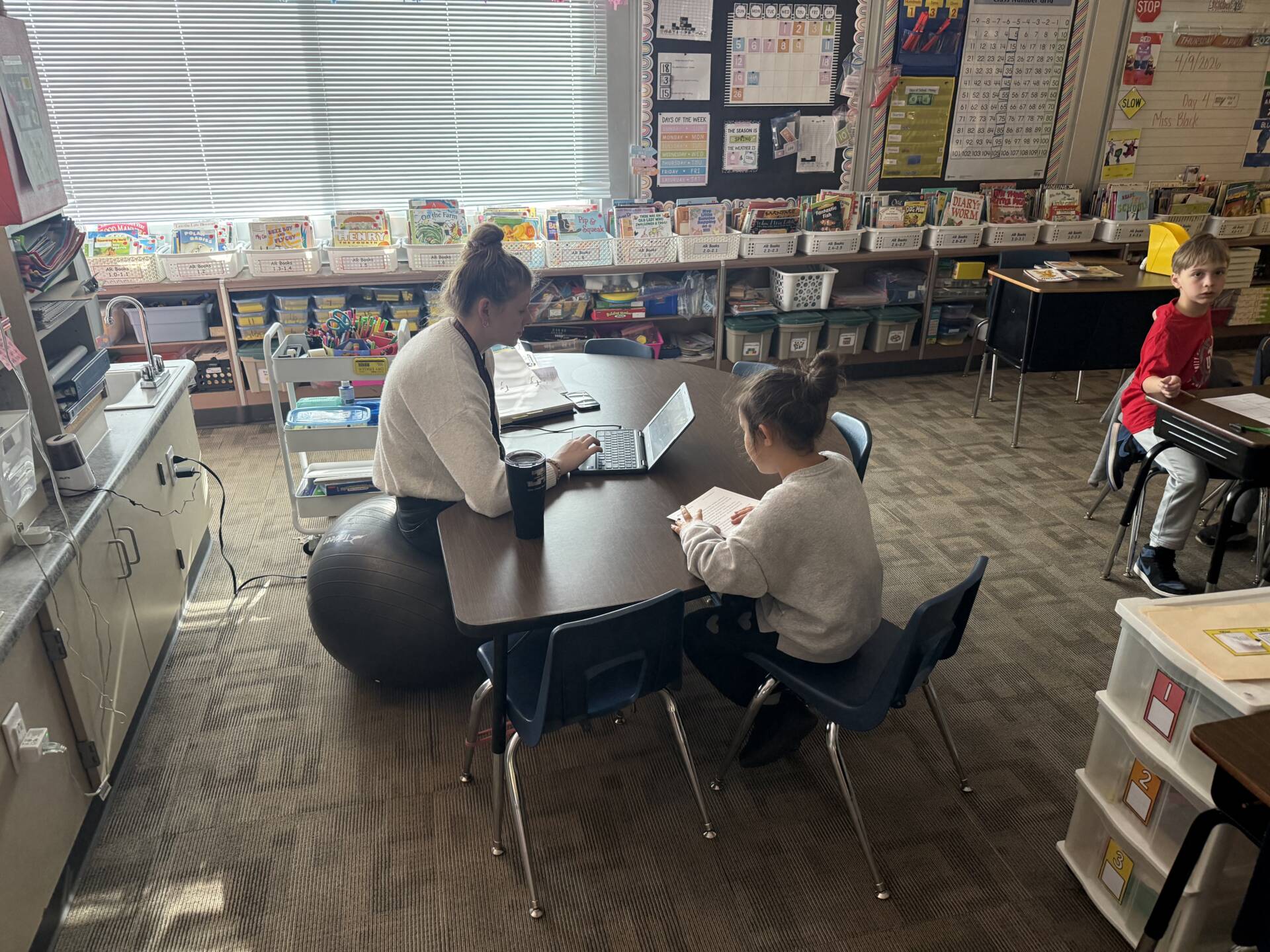Teacher works one-on-one with a student at a table using a laptop, while other students work independently nearby.