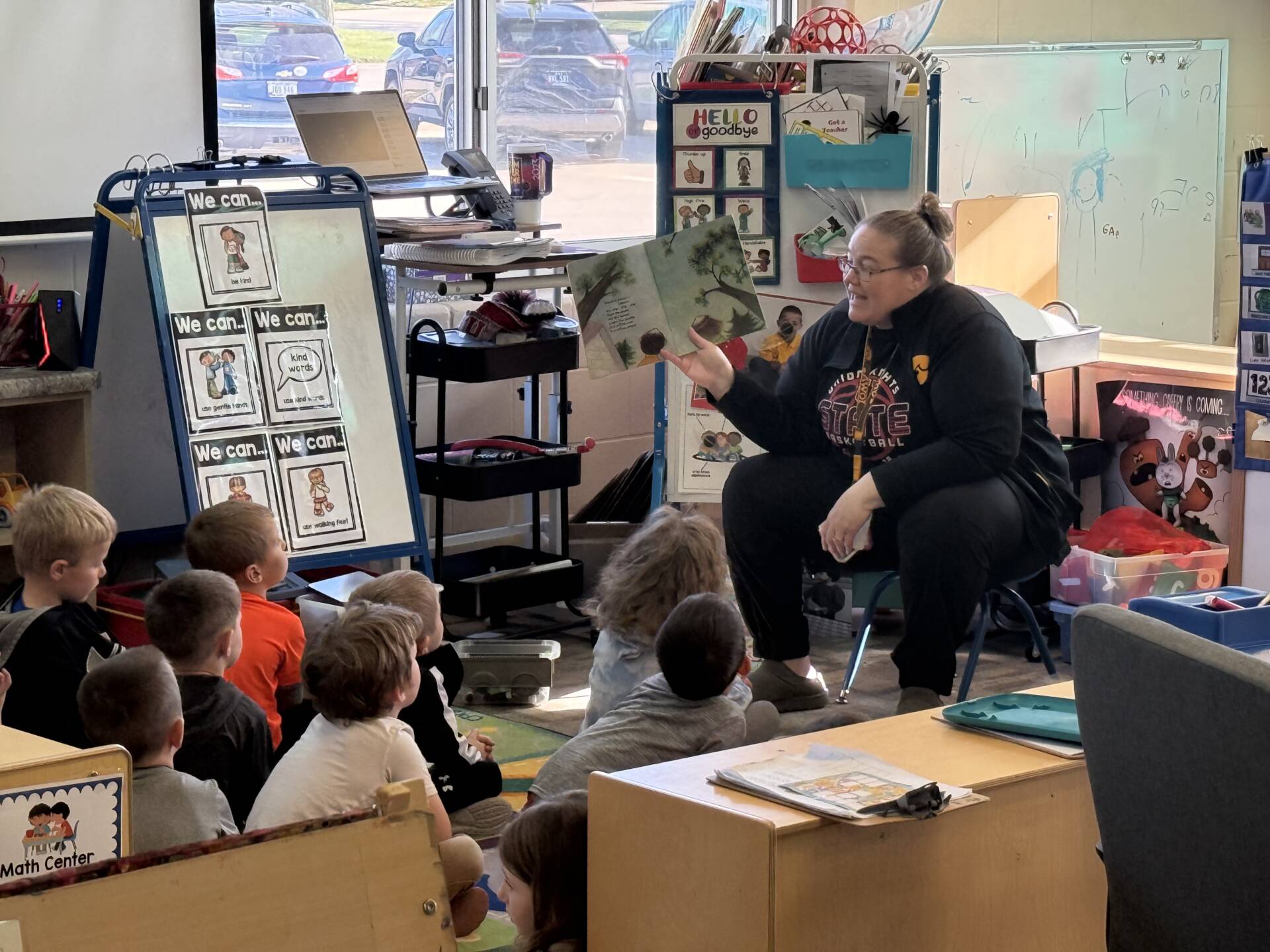 Teacher reads a picture book aloud to young students seated on the floor, who watch attentively in a classroom.