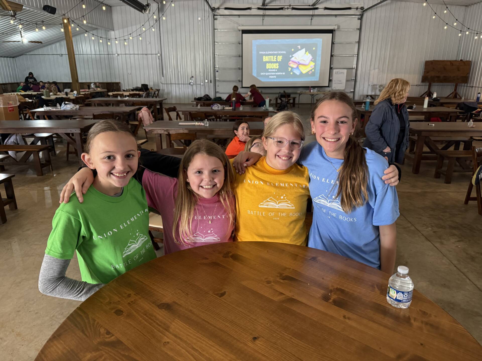 Four girls in colorful shirts stand together smiling at a table, with a “Battle of the Books” screen behind them.