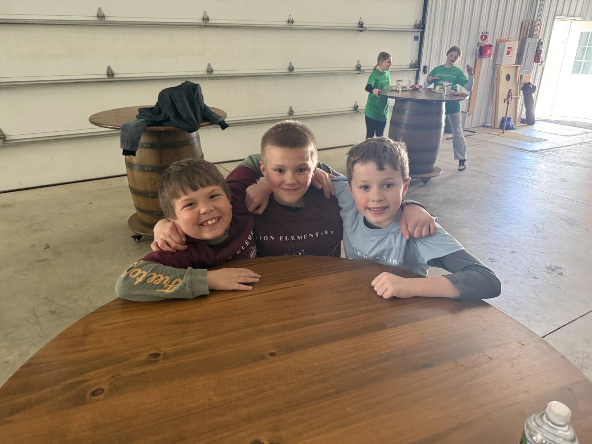Three boys sit at a round wooden table, smiling with arms around each other in a large indoor space.