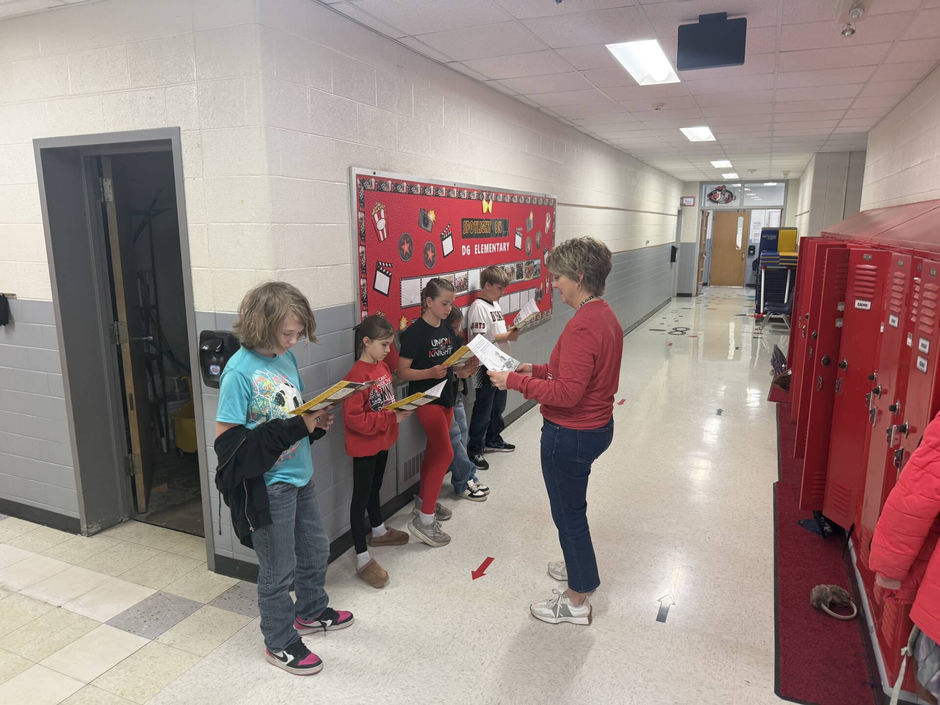 Teacher stands in a hallway guiding students who read papers lined up along the wall near red lockers.