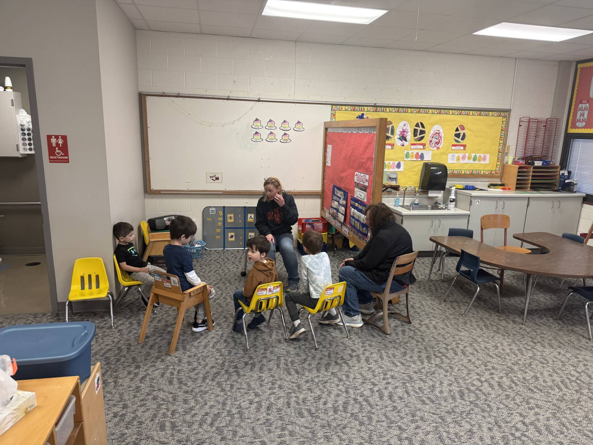 Two adults lead a small group of young children seated in a circle, engaging in a classroom discussion activity.