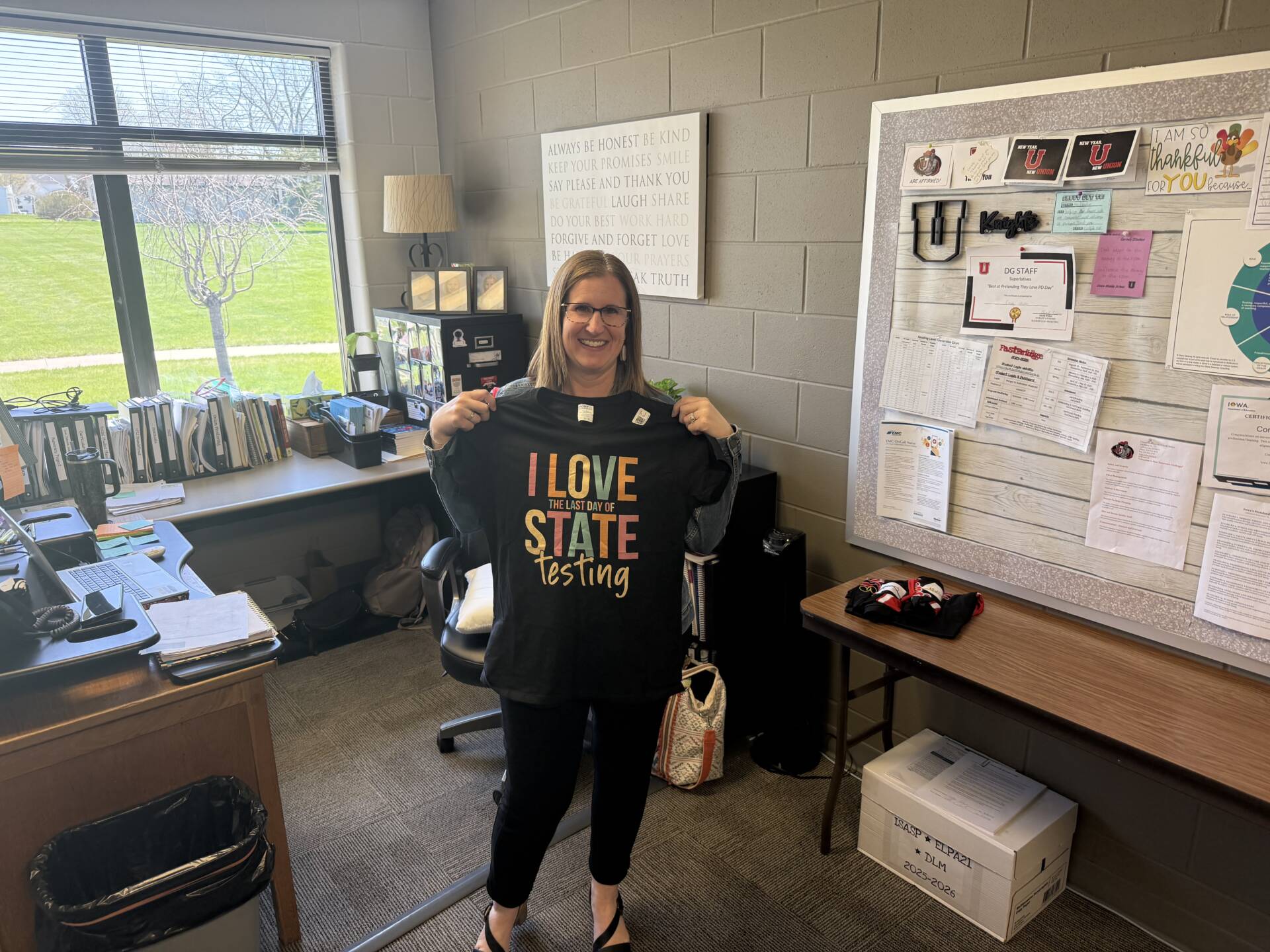 Teacher stands in a classroom holding a shirt that reads “I love the last day of state testing,” smiling.