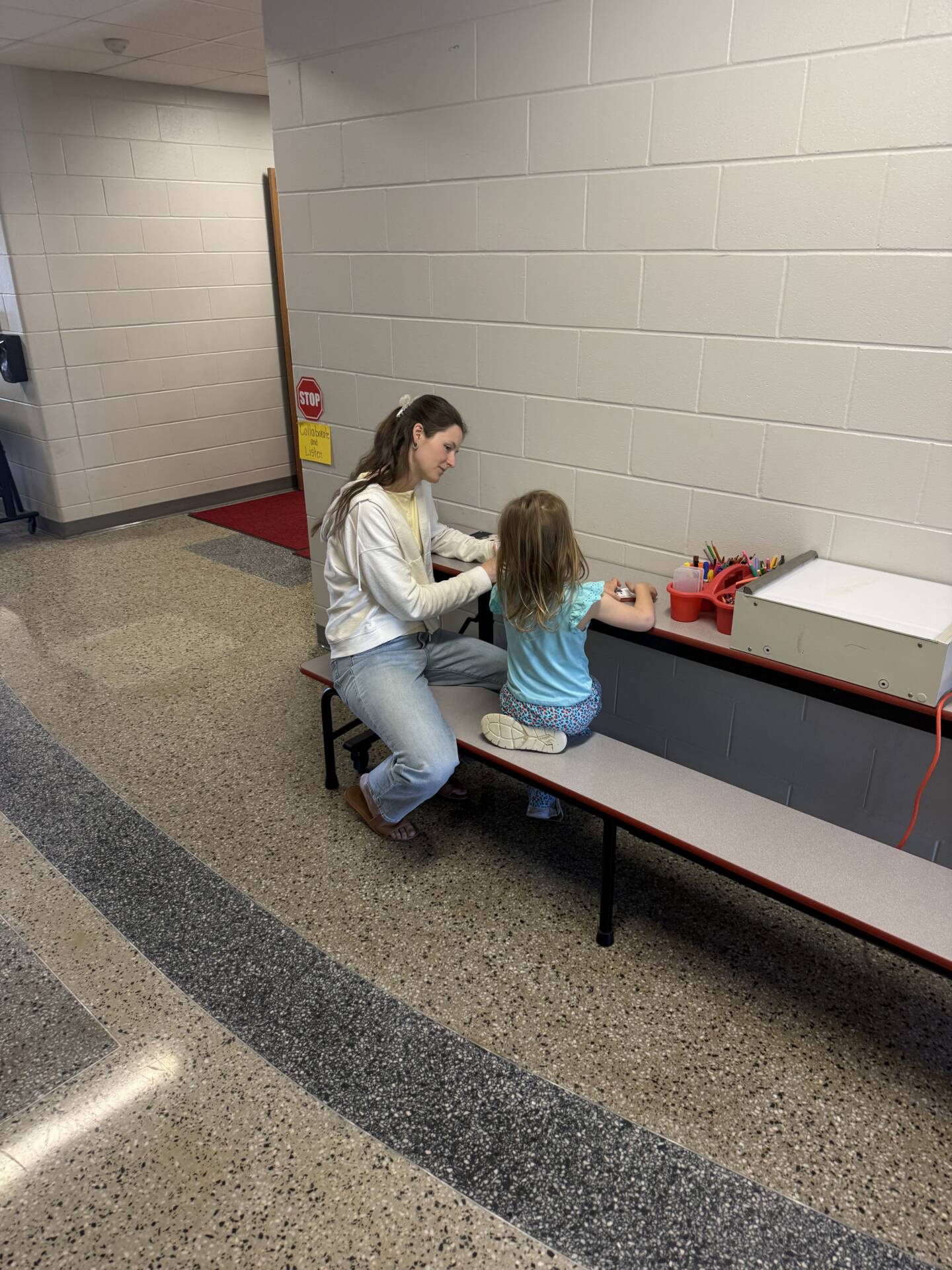 Adult sits beside a young child at a bench, helping them draw with markers in a quiet hallway workspace.