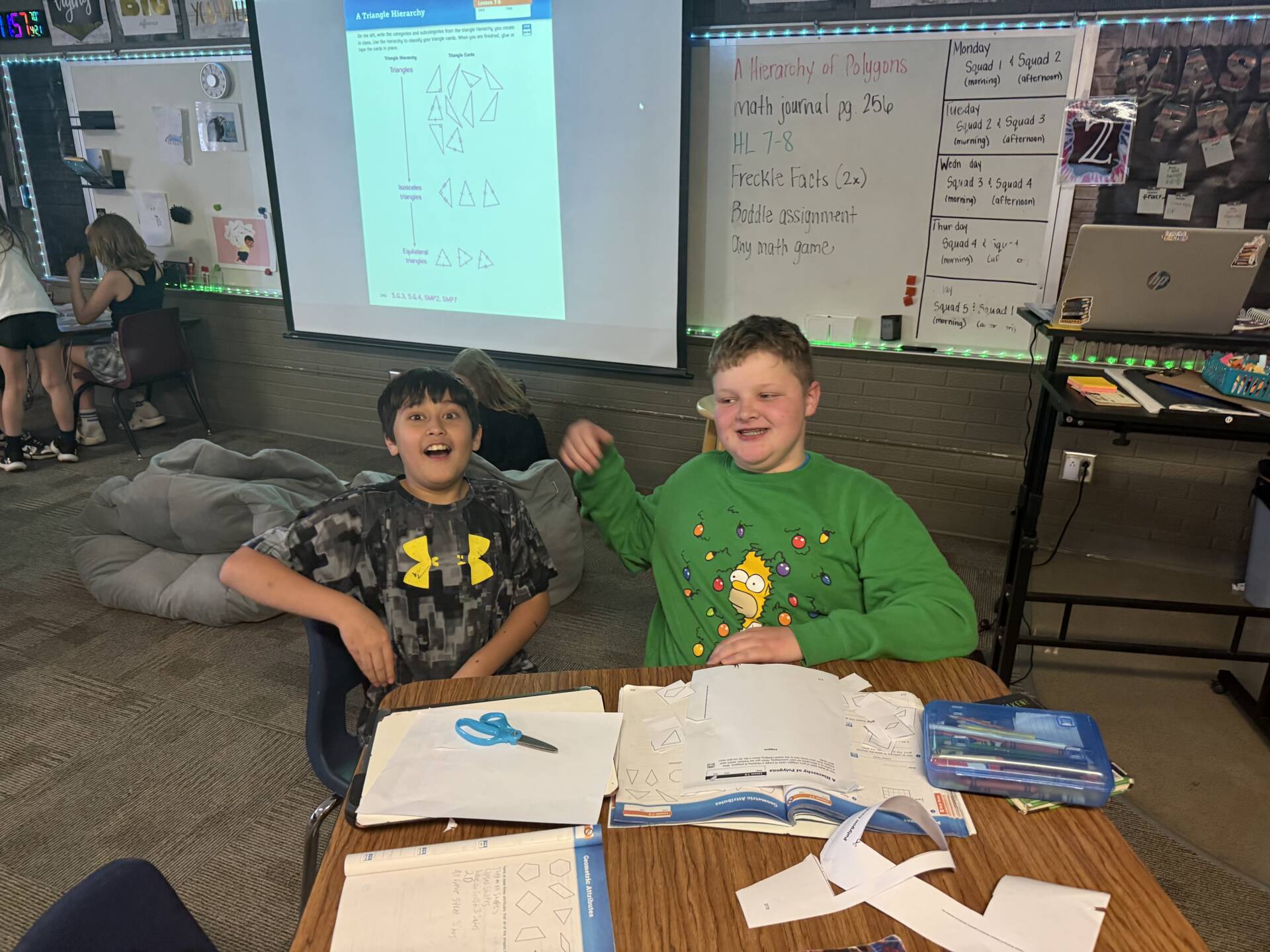Two students sit at a desk smiling during a math activity with cut paper shapes and open workboo