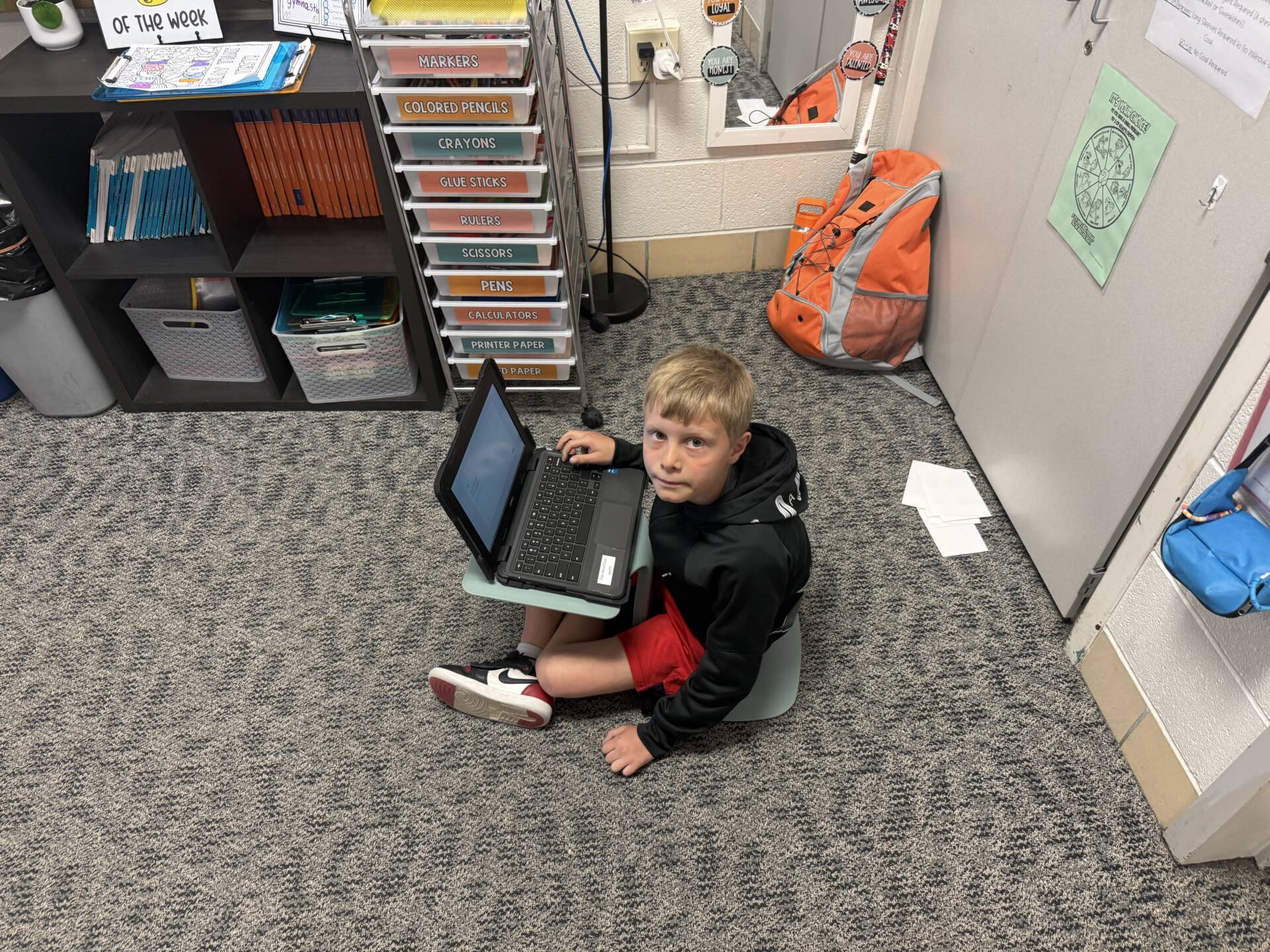 Student sits on the floor using a laptop on a small desk, working independently in a classroom corner.