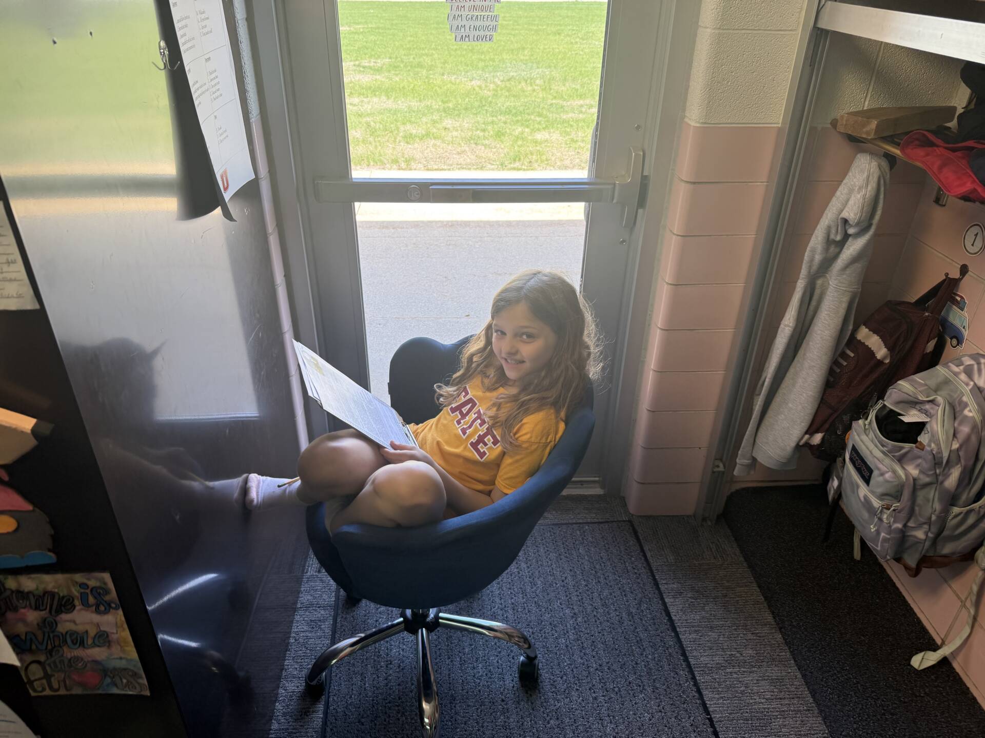Student sits curled in a chair by a window, smiling while reading a paper in a quiet classroom corner.