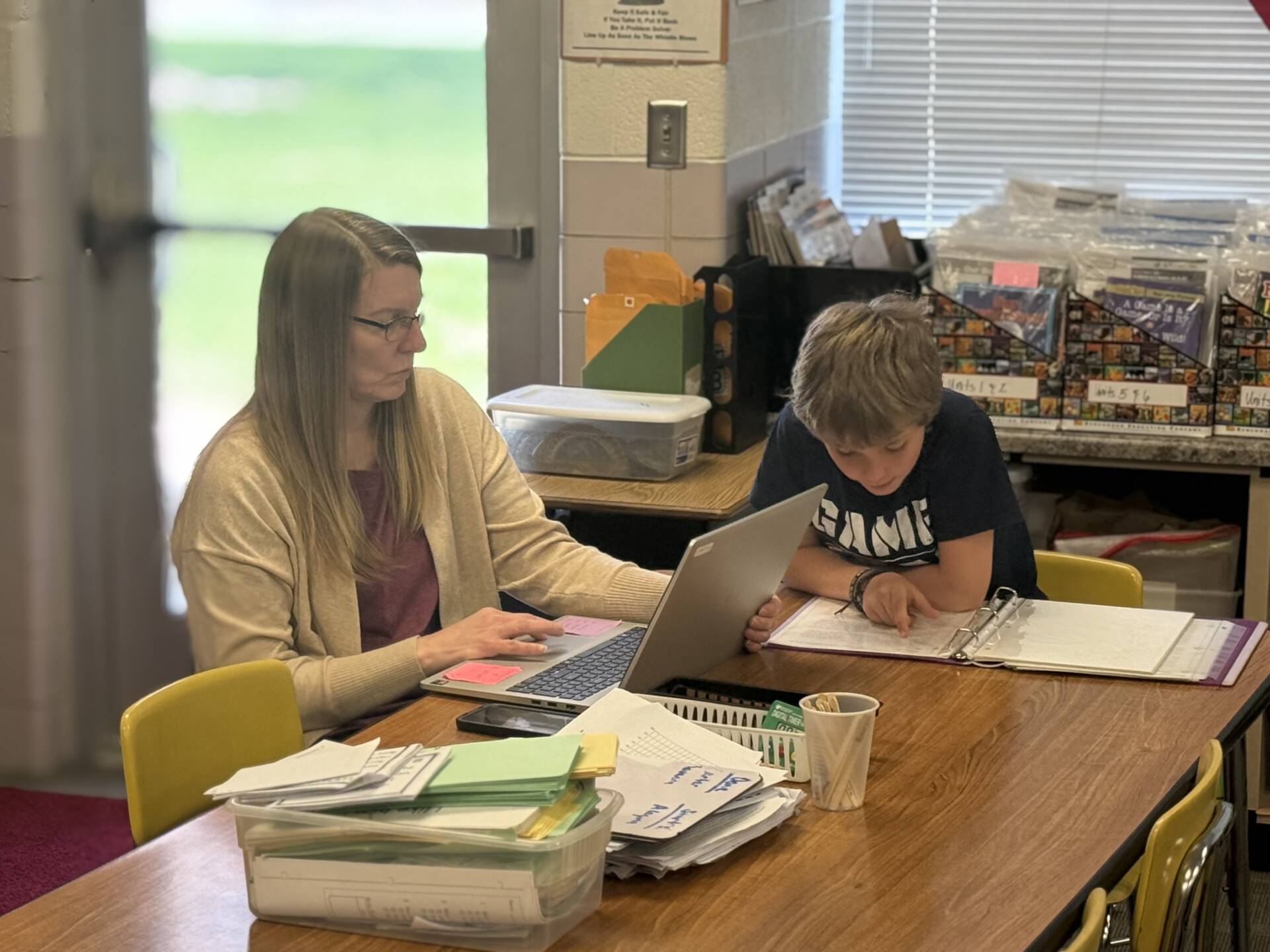Teacher works on a laptop beside a student who reads from a binder during small group instruction.