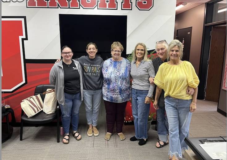 Seven women stand together in a school office area, smiling in front of a “Knights” sign and a large screen.