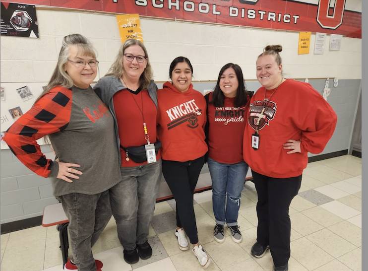 Five staff members stand together in a school hallway wearing red and black “Knights” apparel, smiling with arms around each other.