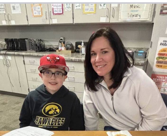 Adult and student sit side by side at a desk, smiling in a classroom with cabinets and supplies in the background.