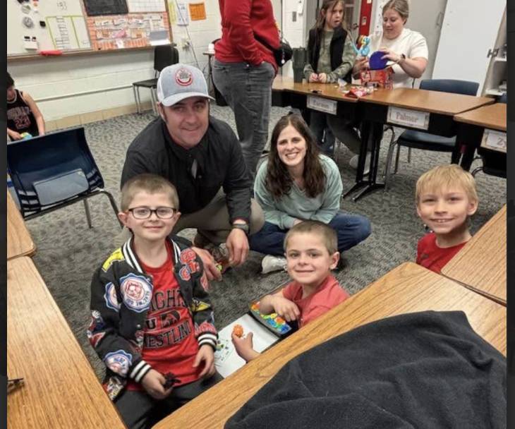 Two adults and three young students smile and sit together on the floor, working with small building pieces in a classroom.