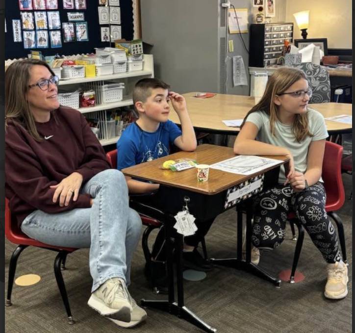 Adult and two students sit at desks, listening attentively during a classroom activity with papers and supplies on the table.