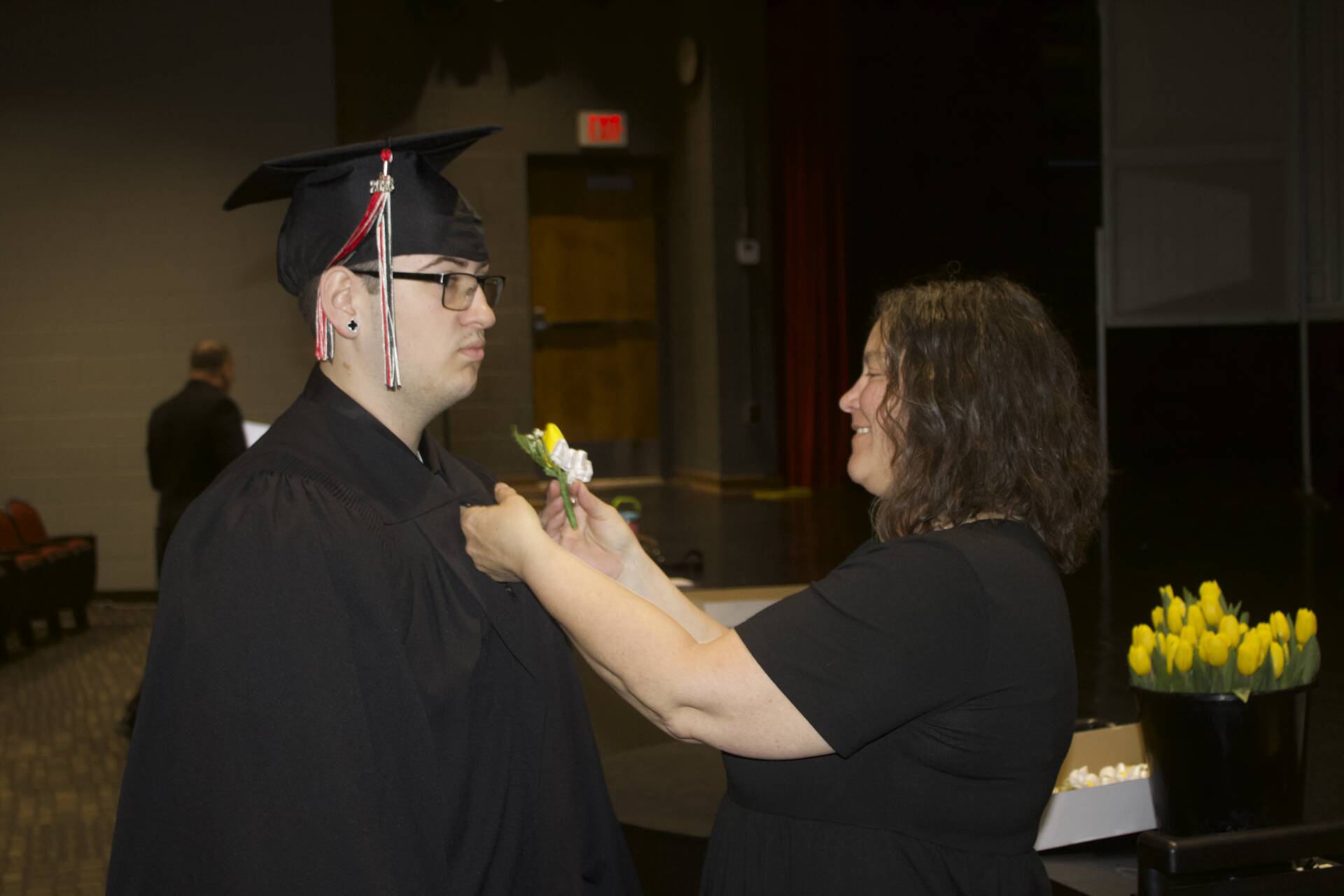 Adult pins a white flower boutonniere onto a graduate’s black gown before the ceremony begins.
