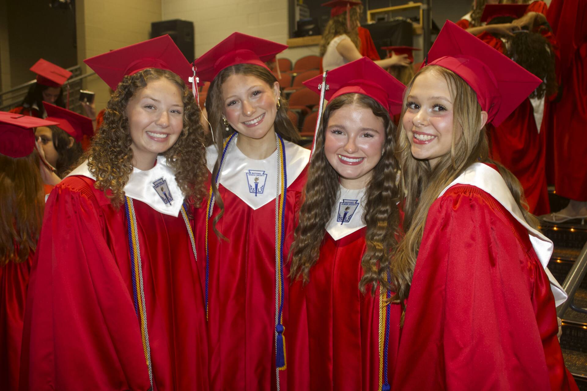 Four smiling graduates in red caps and gowns pose together near auditorium seating before graduation.