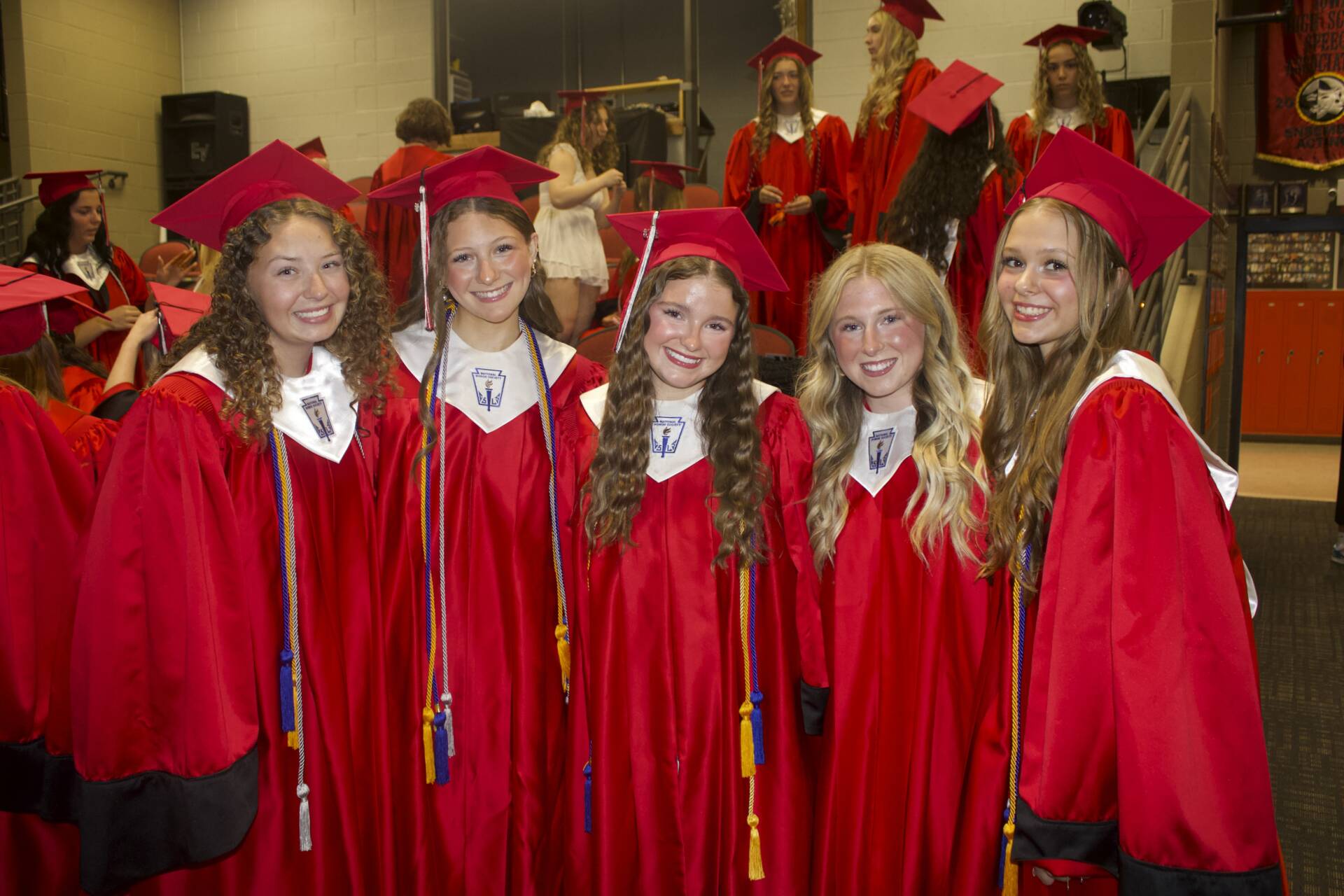 Group of graduates in red caps and gowns gather backstage, smiling before commencement begins.