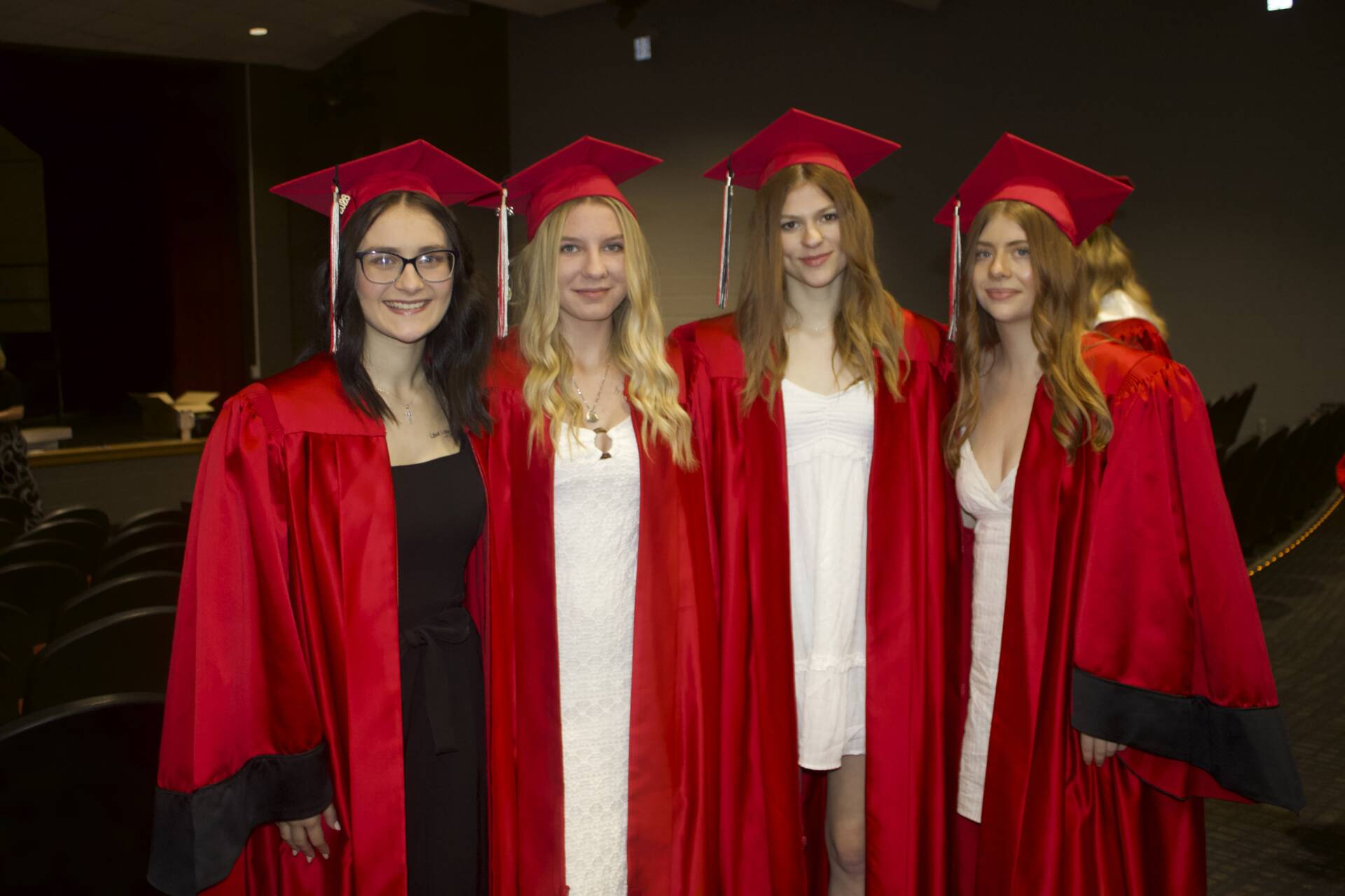 Four graduates in red caps and gowns smile together in an auditorium before graduation.