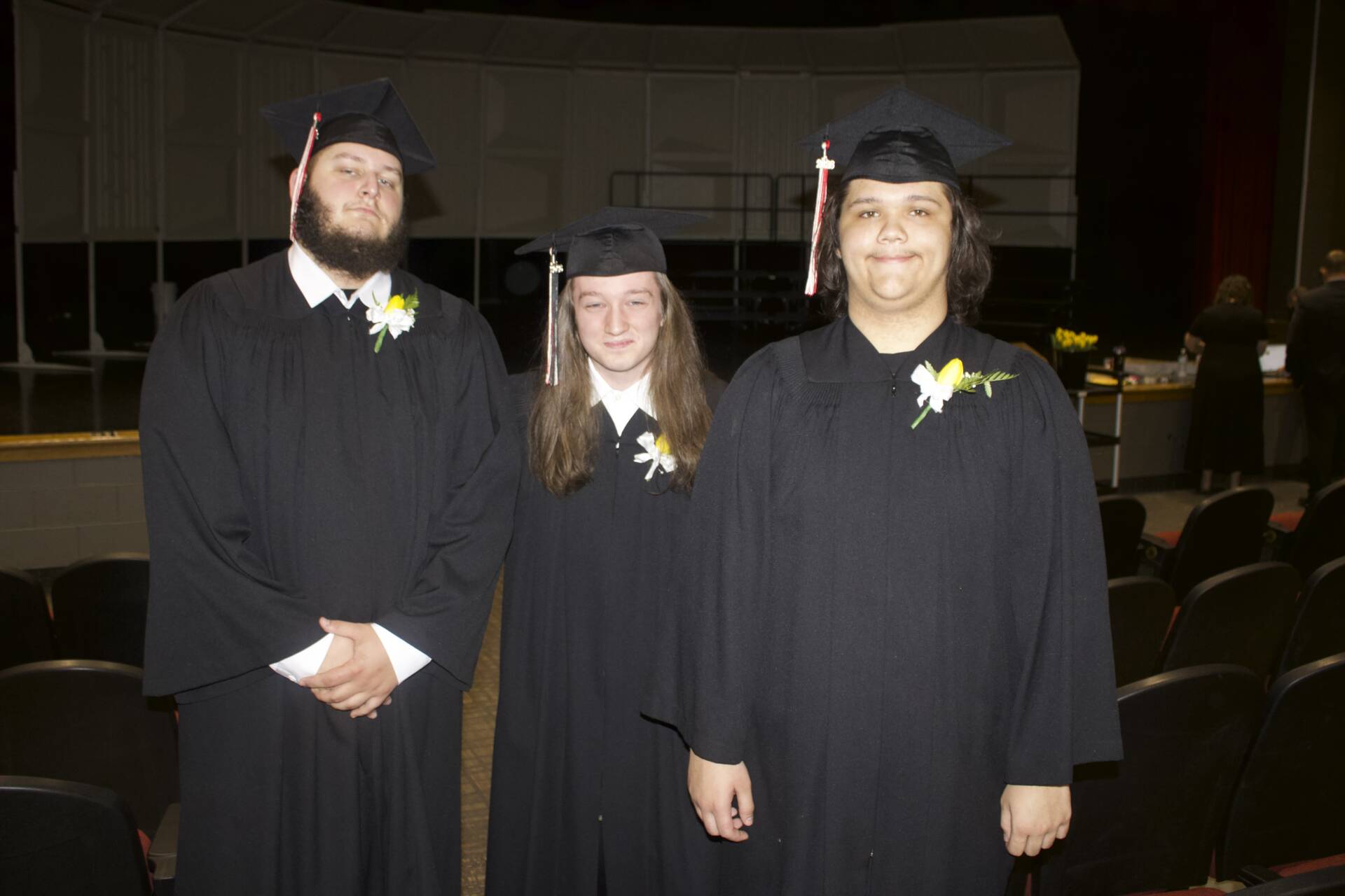 Three graduates in black caps and gowns stand together onstage before a graduation ceremony.