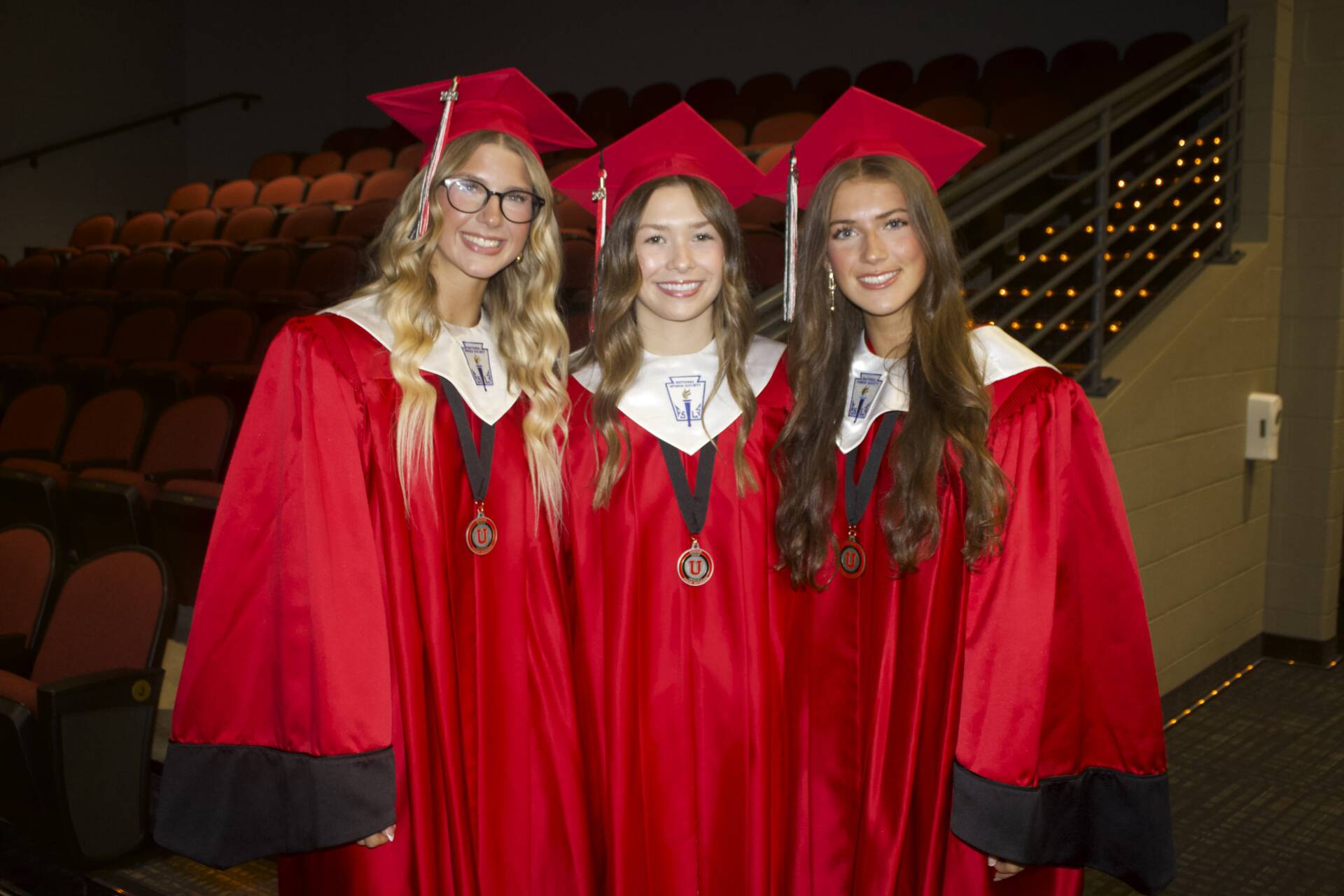 Three smiling graduates in red caps and gowns pose together in an auditorium before commencement.