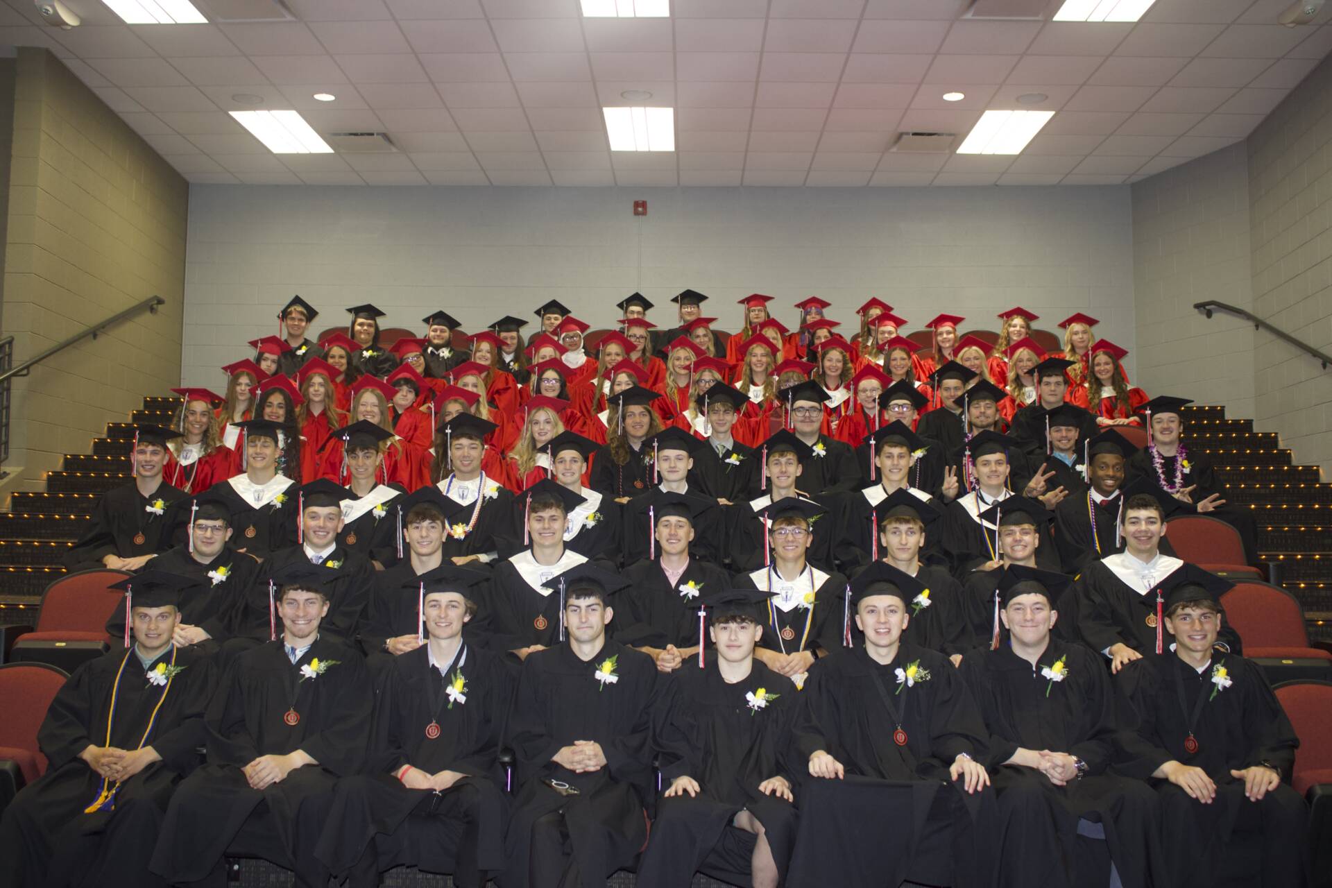 Graduating class in red and black caps and gowns seated together in an auditorium for a group graduation photo.
