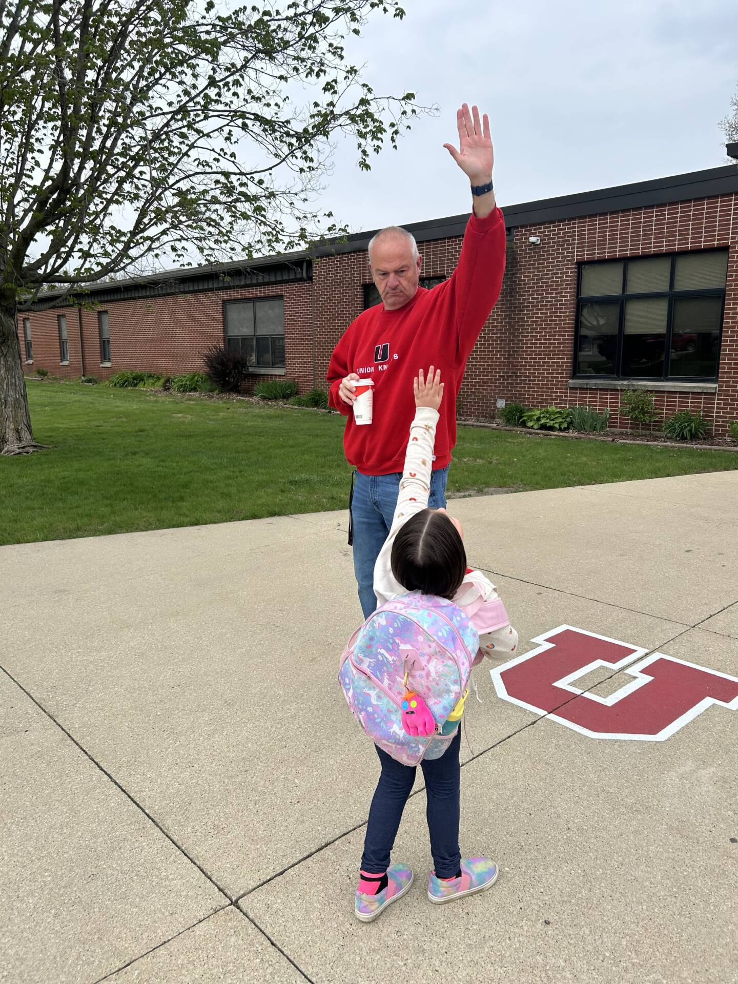 School staff member in a red sweatshirt high-fives a young student with a colorful backpack outside the school building.