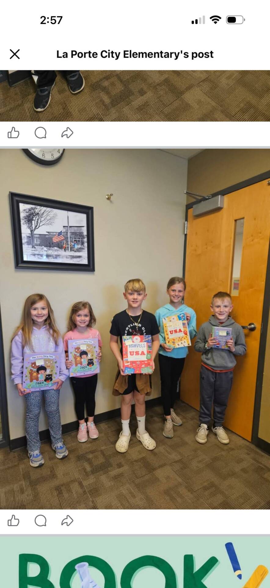 Five elementary students stand indoors holding books, smiling near a classroom door and wall clock.