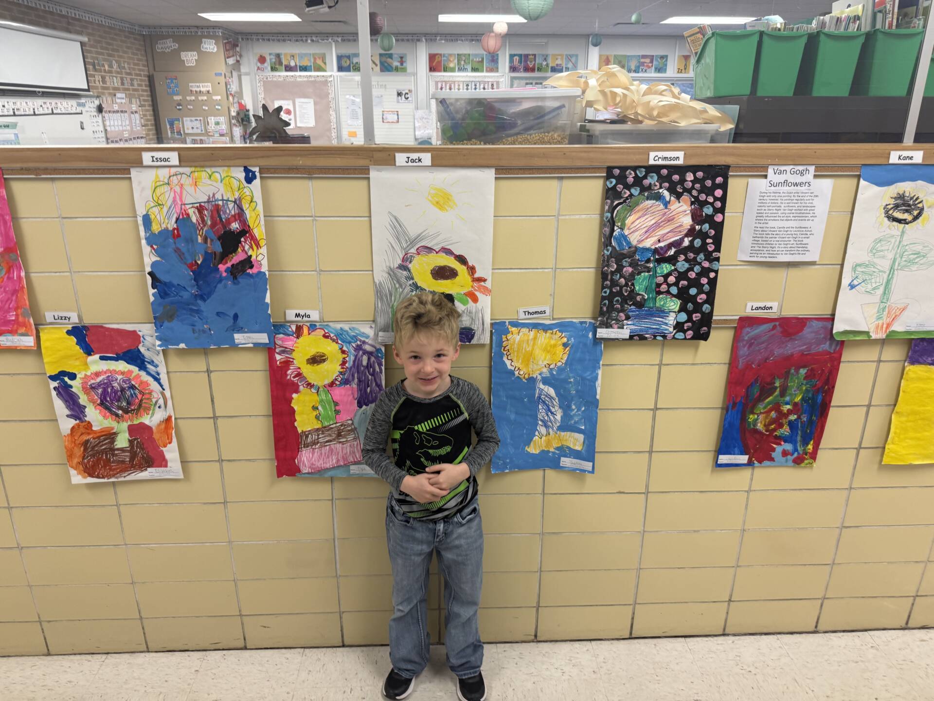 Young boy stands in hallway in front of a display of bright student paintings, smiling with hands clasped.