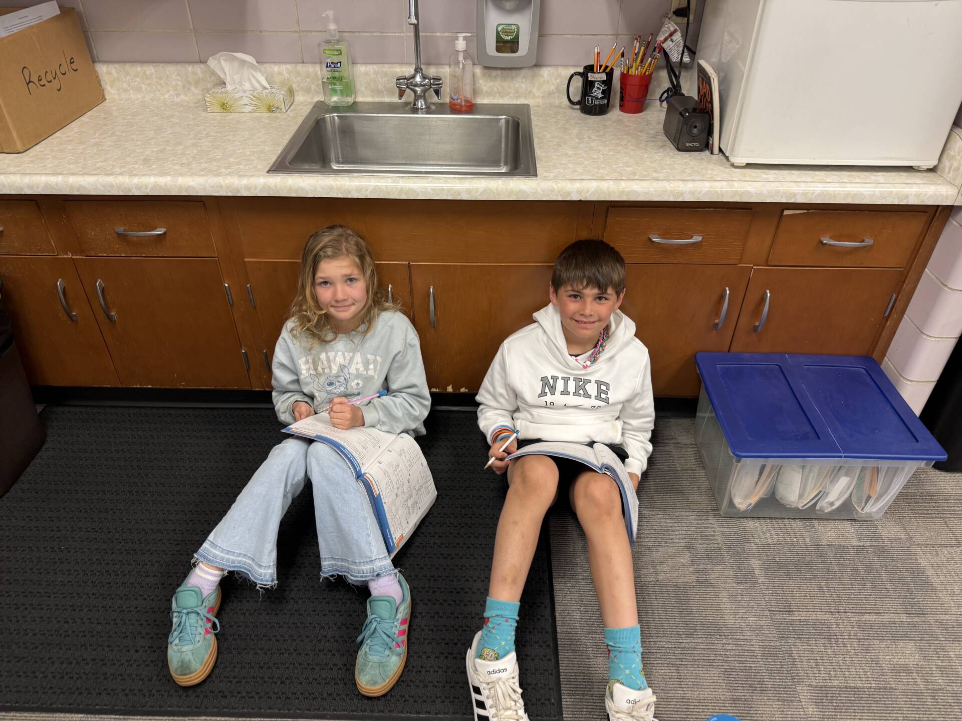 Two children sit on the floor by cabinets, writing in workbooks with pencils during a classroom activity.