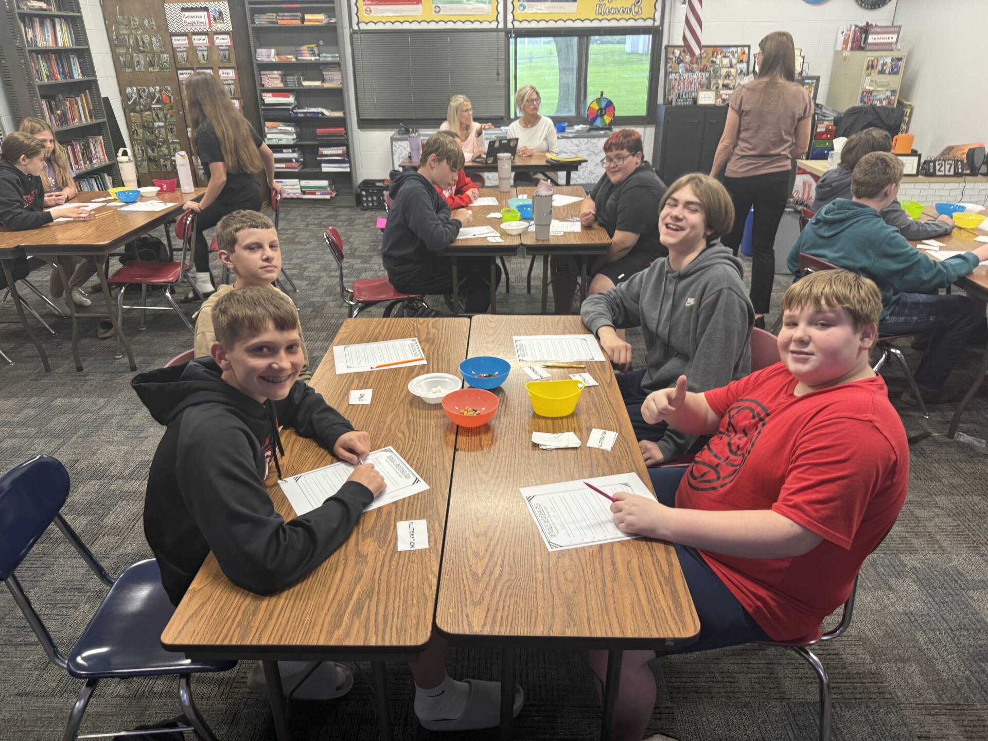 Middle school students sit at desks in a classroom, working on papers with snacks, chatting and smiling.