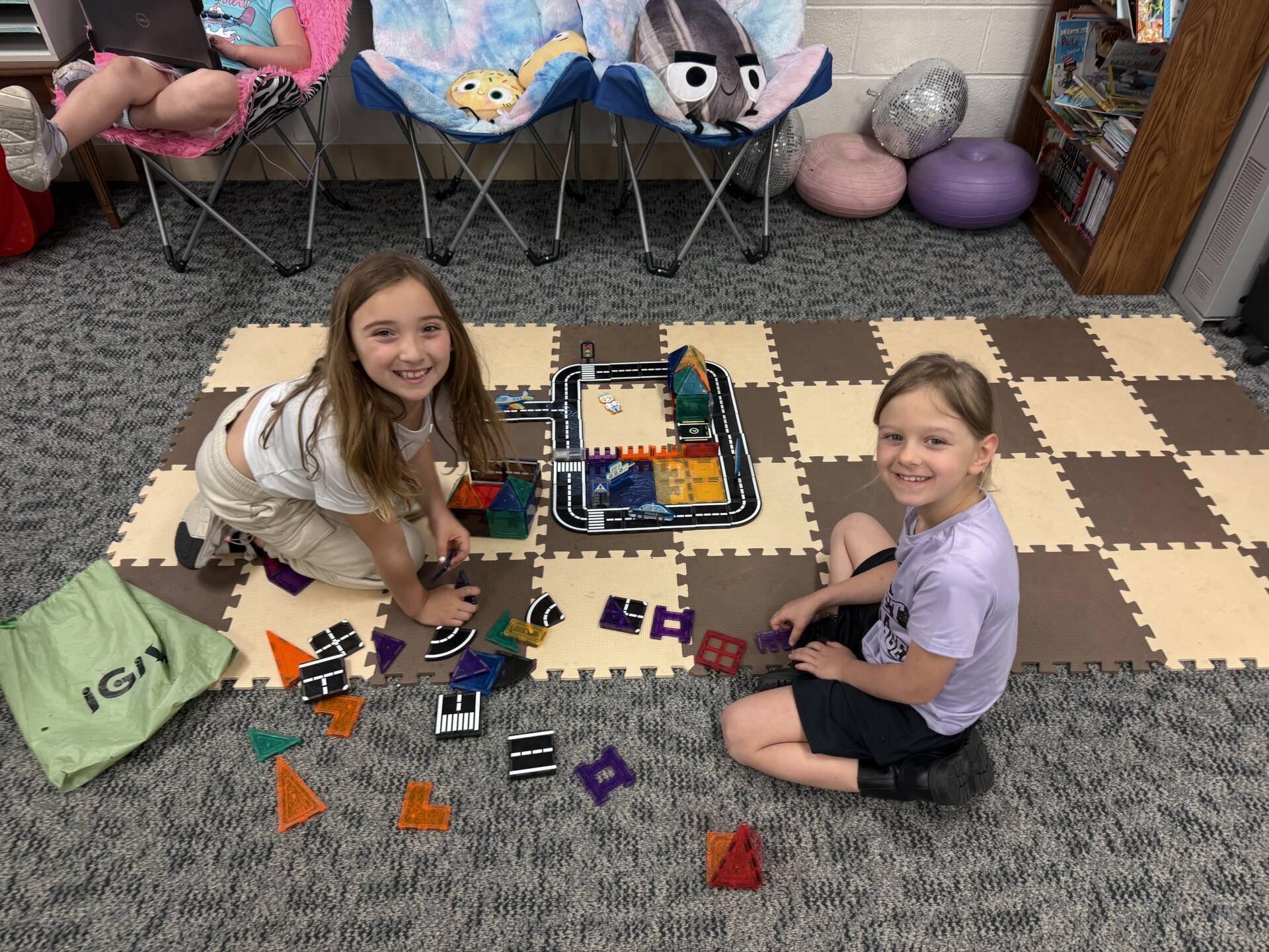 Two young girls sit on a classroom mat building with colorful magnetic tiles and a small road set, smiling at camera.