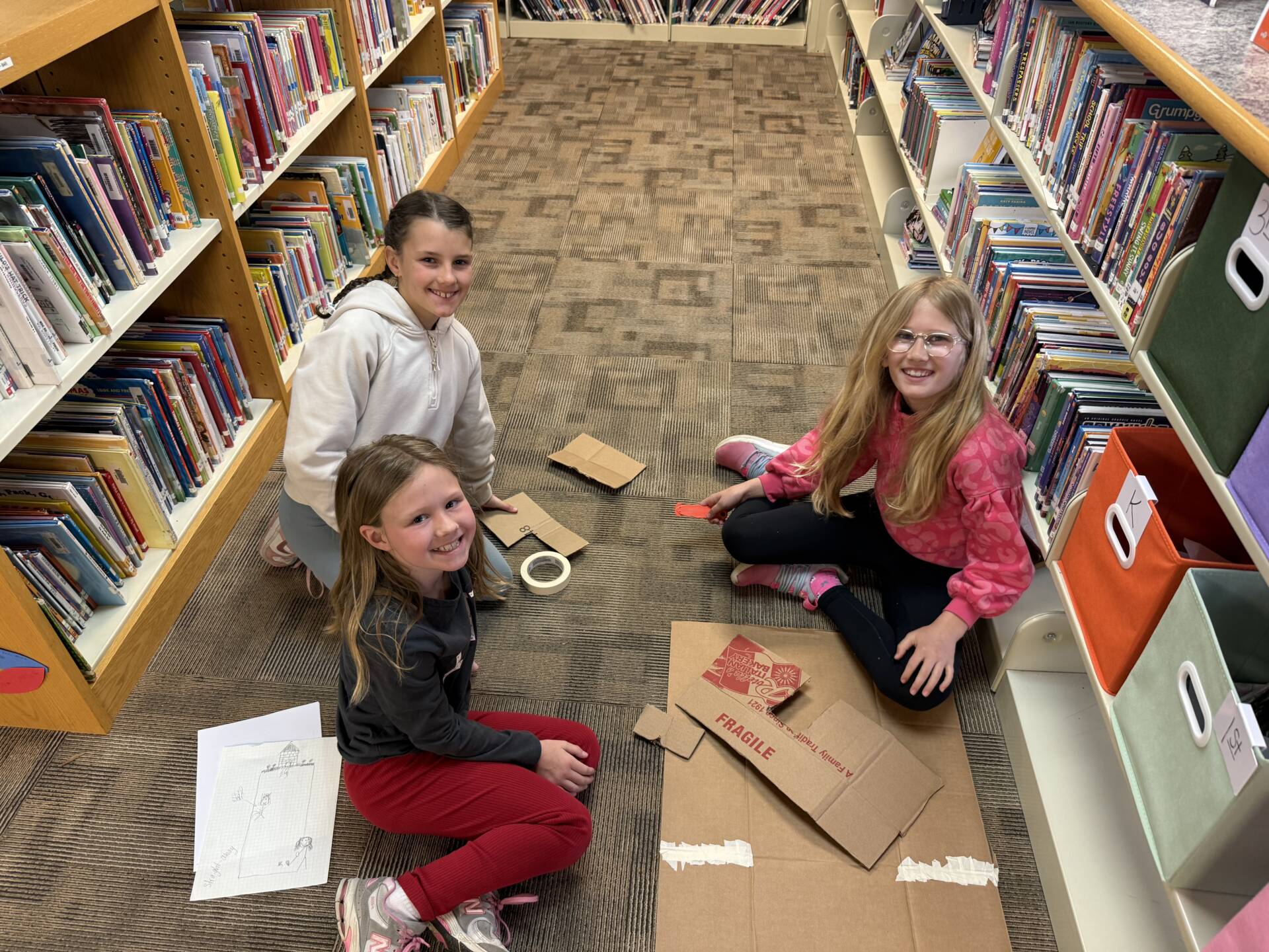 Three elementary students sit on a library floor building cardboard creations between bookshelves.