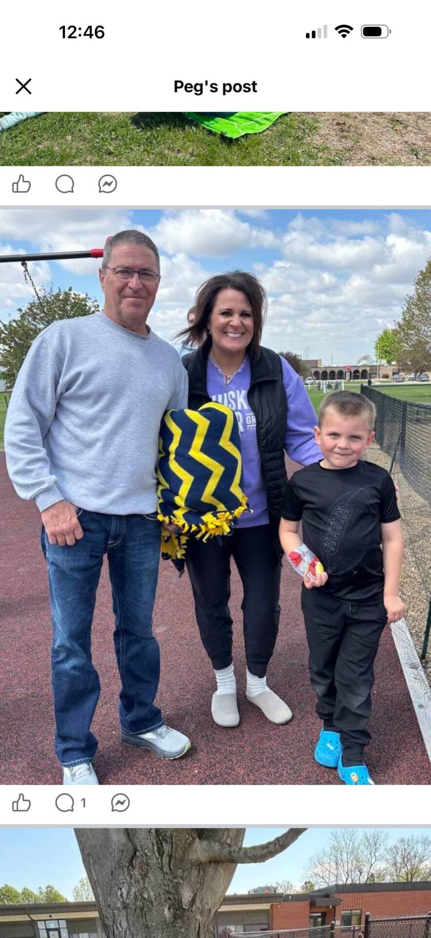Family of three smiles outdoors near a playground and walking track on a cloudy day.