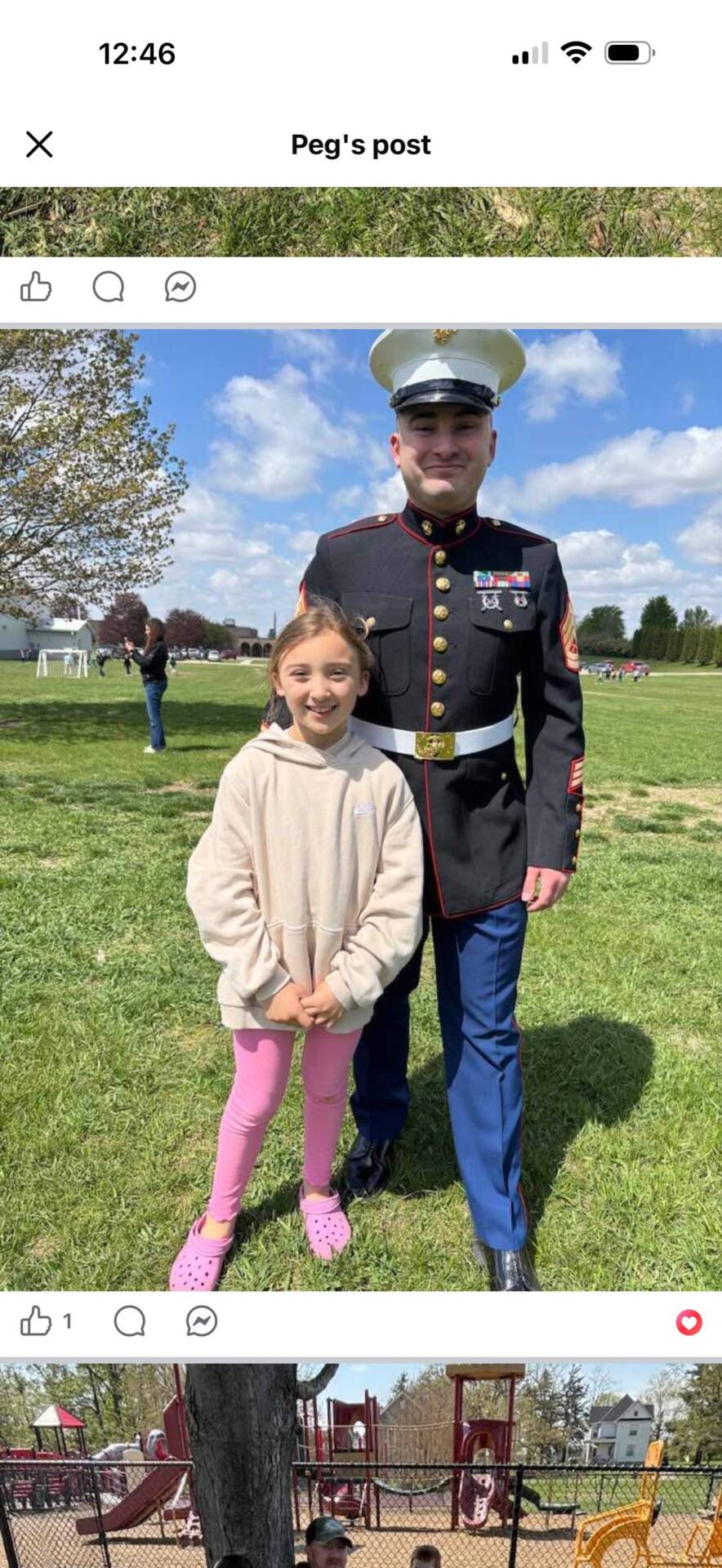 Young girl stands beside a Marine in dress uniform on a grassy field under a blue sky.