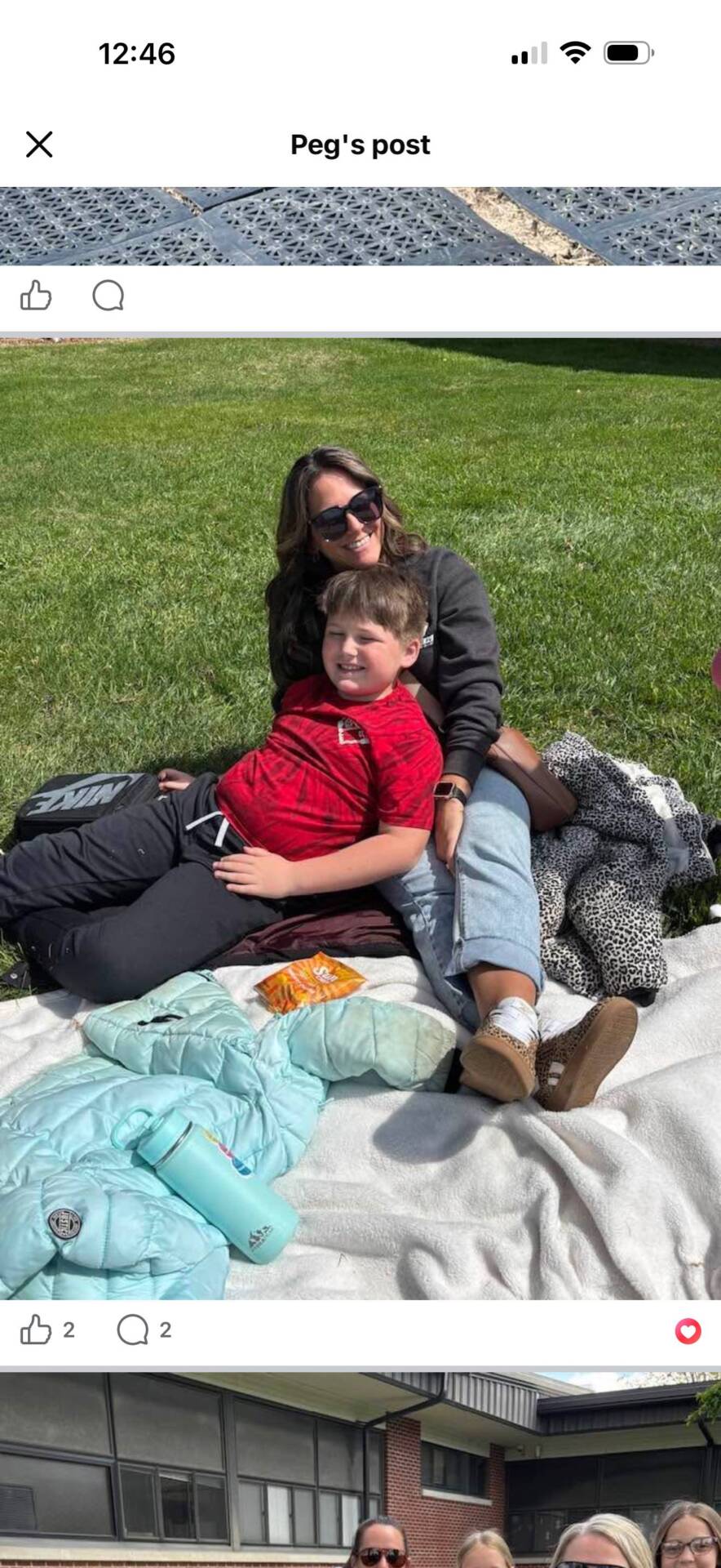 Adult and child sit together on a blanket in the grass during an outdoor school or community event.