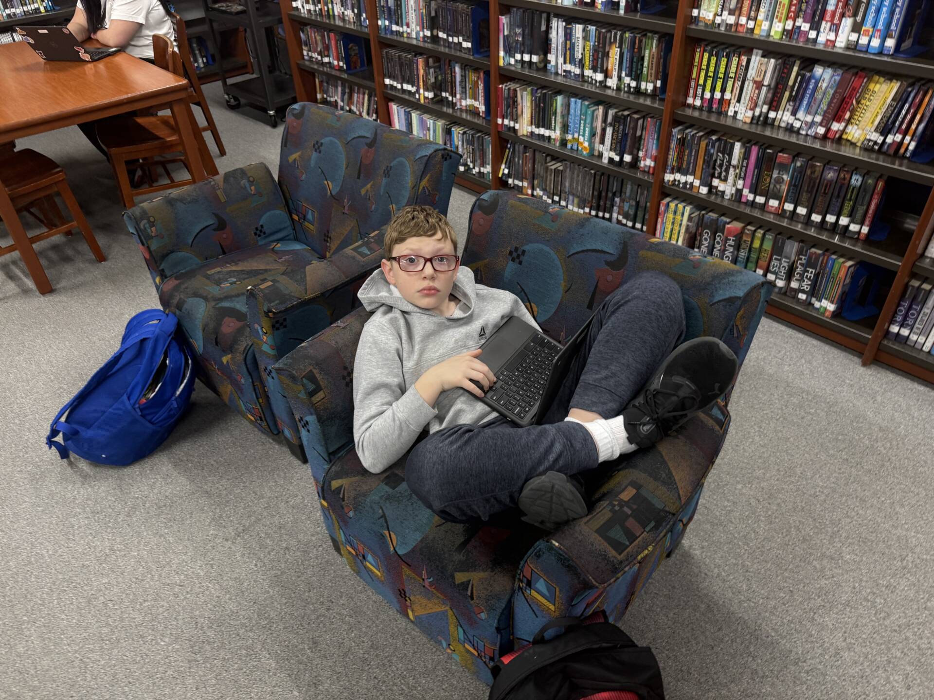 Student lounges in a library armchair using a tablet computer, surrounded by bookshelves and study tables.