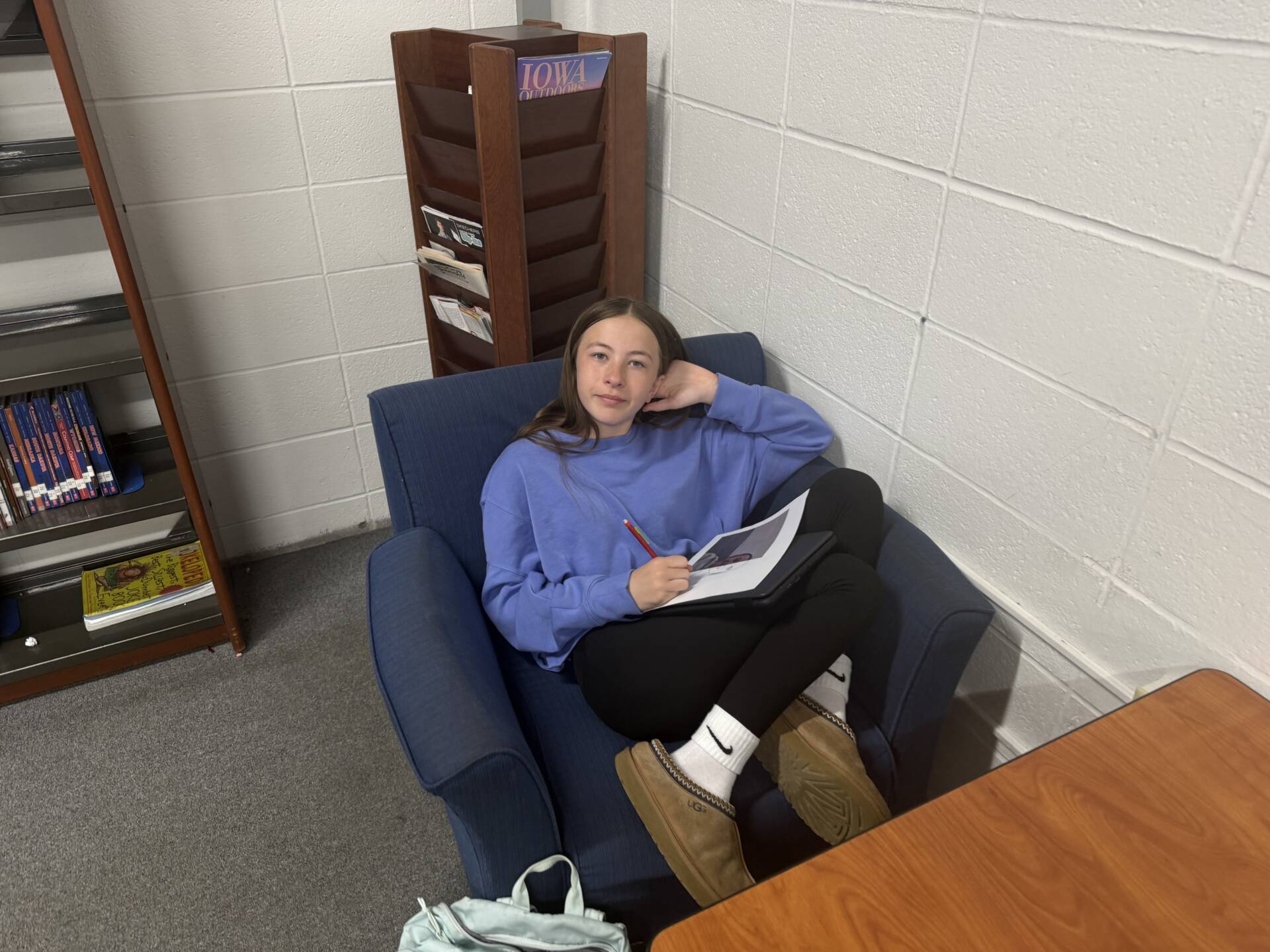 Student relaxes in a library chair while writing on a worksheet with a pencil and tablet on her lap.