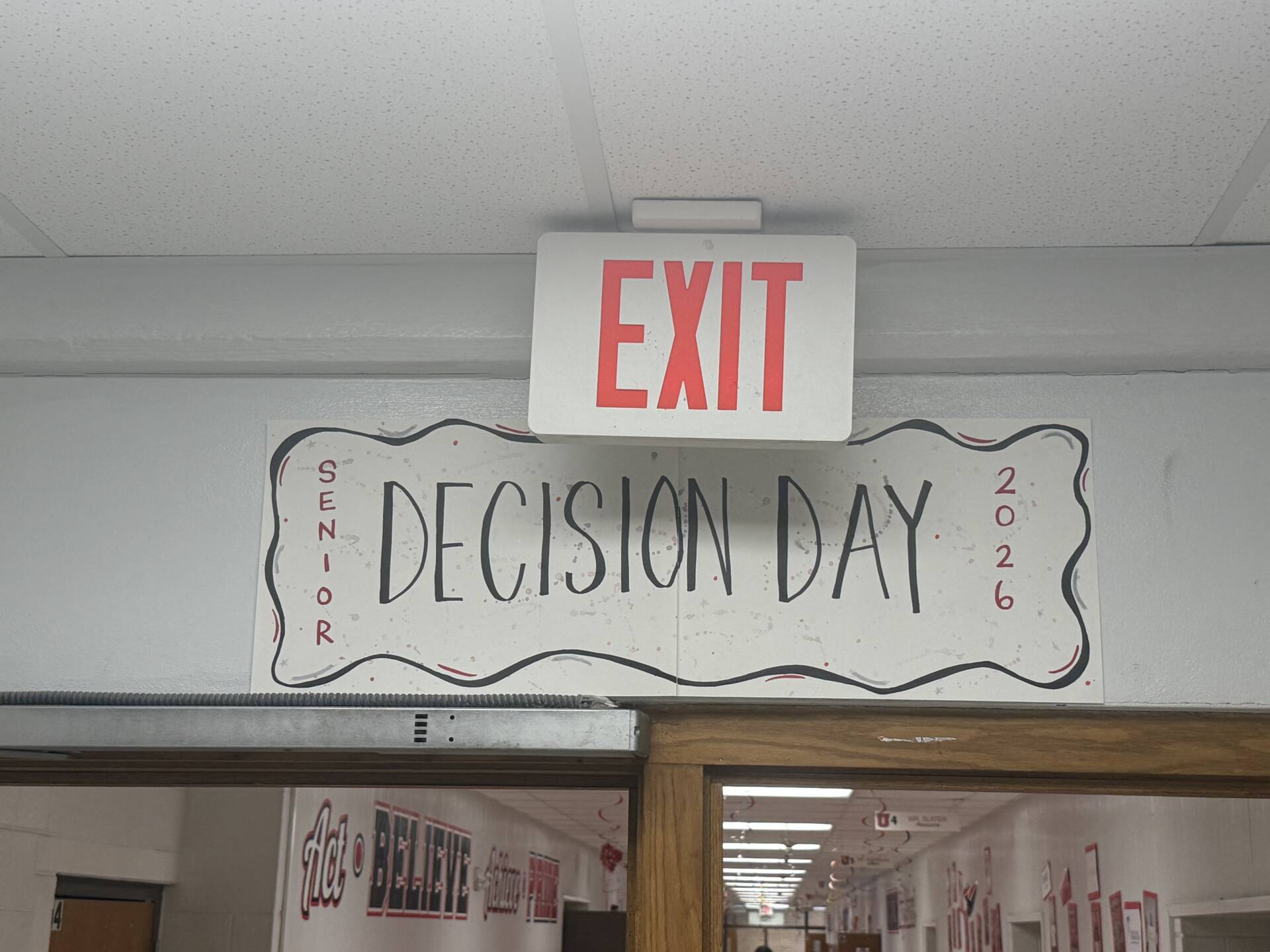 Hallway sign reading “Senior Decision Day 2026” hangs beneath a glowing exit sign above school doors.
