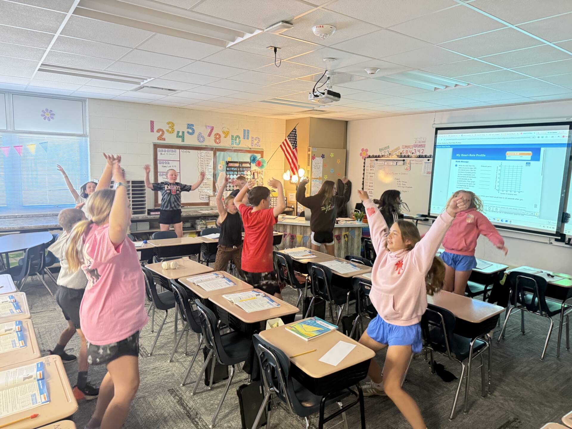 Students stand beside desks with raised arms during an energetic classroom activity in an elementary classroom.