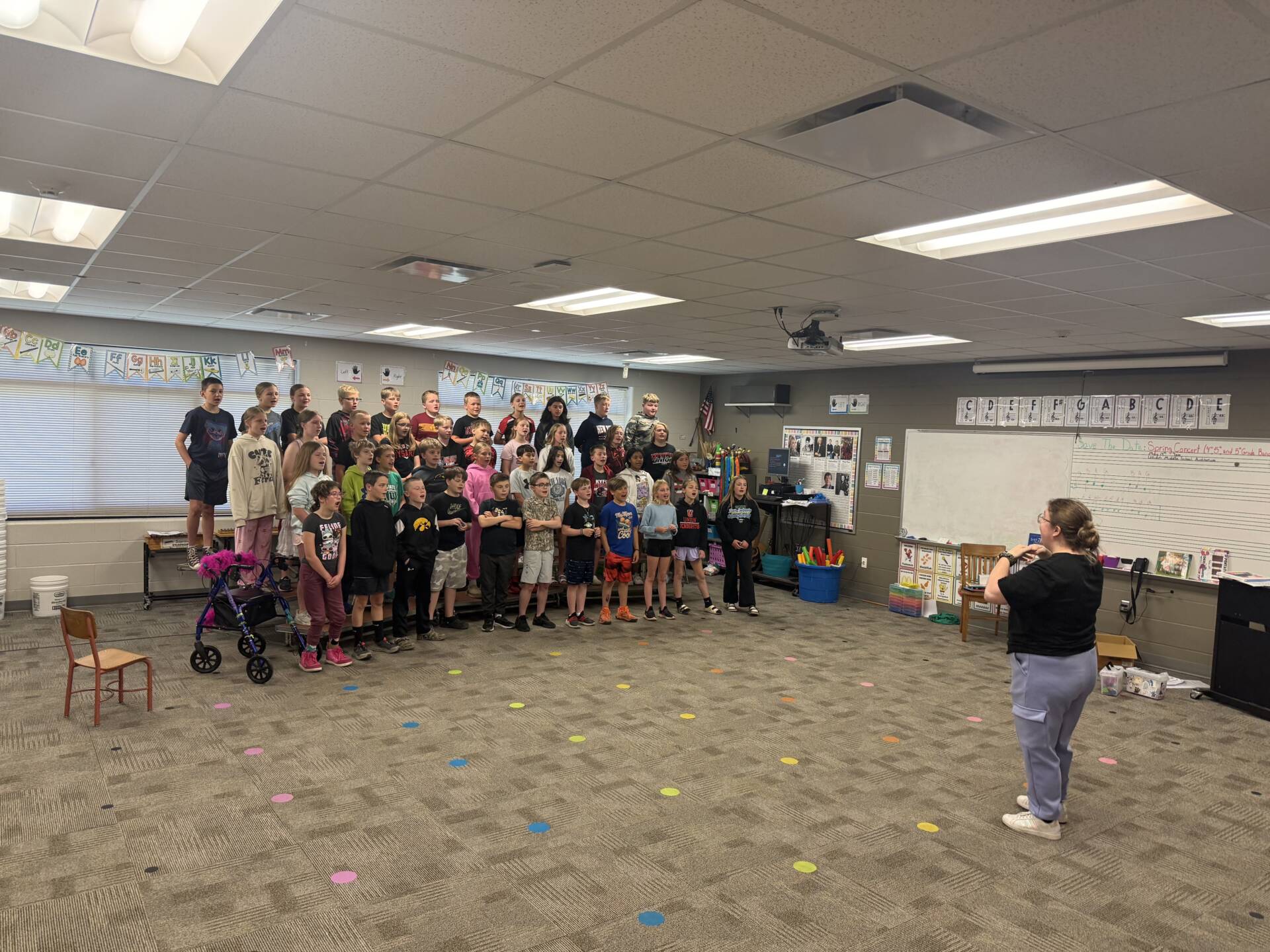 Elementary students sing on risers while a teacher directs rehearsal in a brightly lit music classroom.
