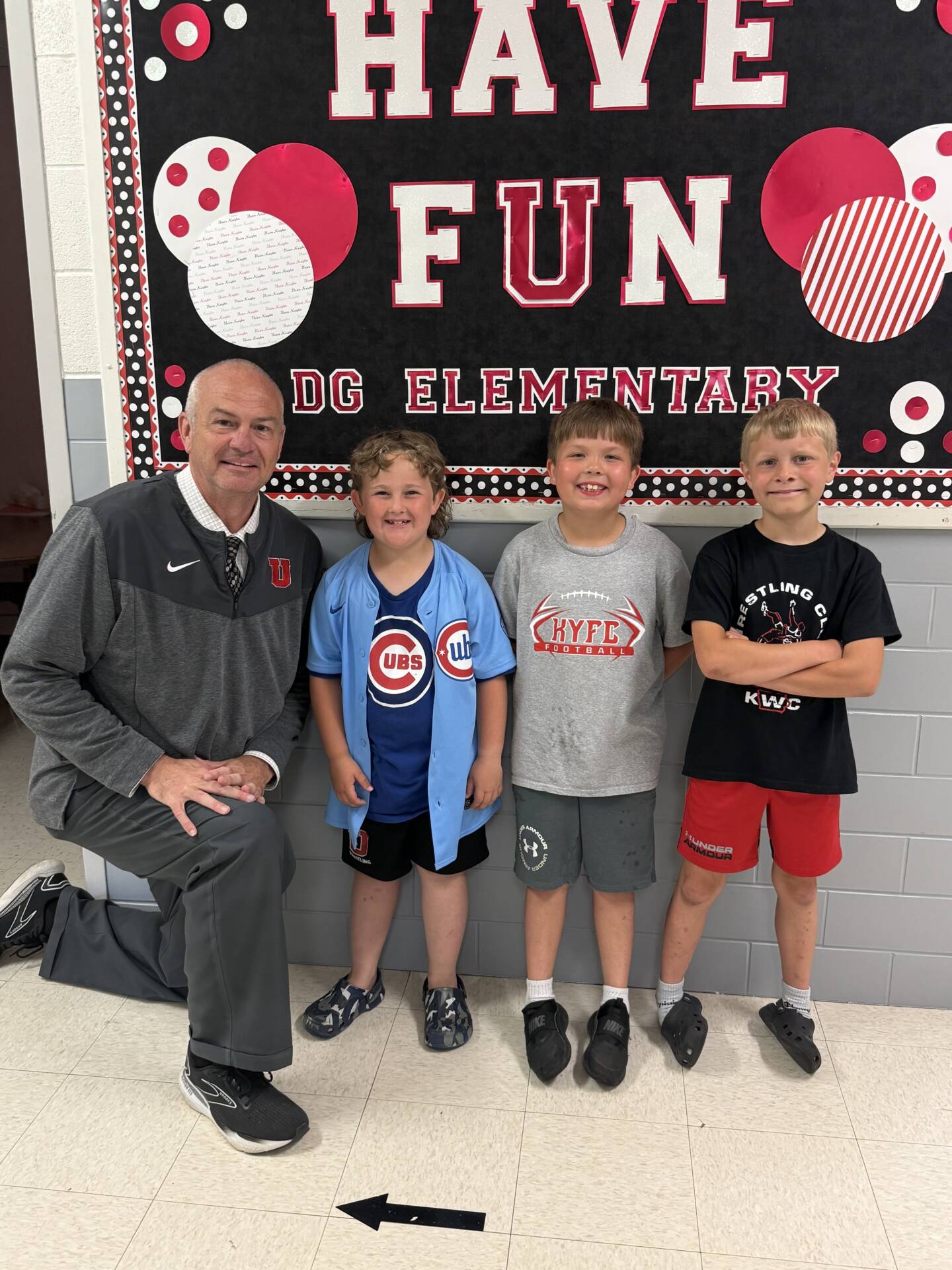 School staff member kneels beside three elementary students smiling in front of a “Have Fun” hallway display.