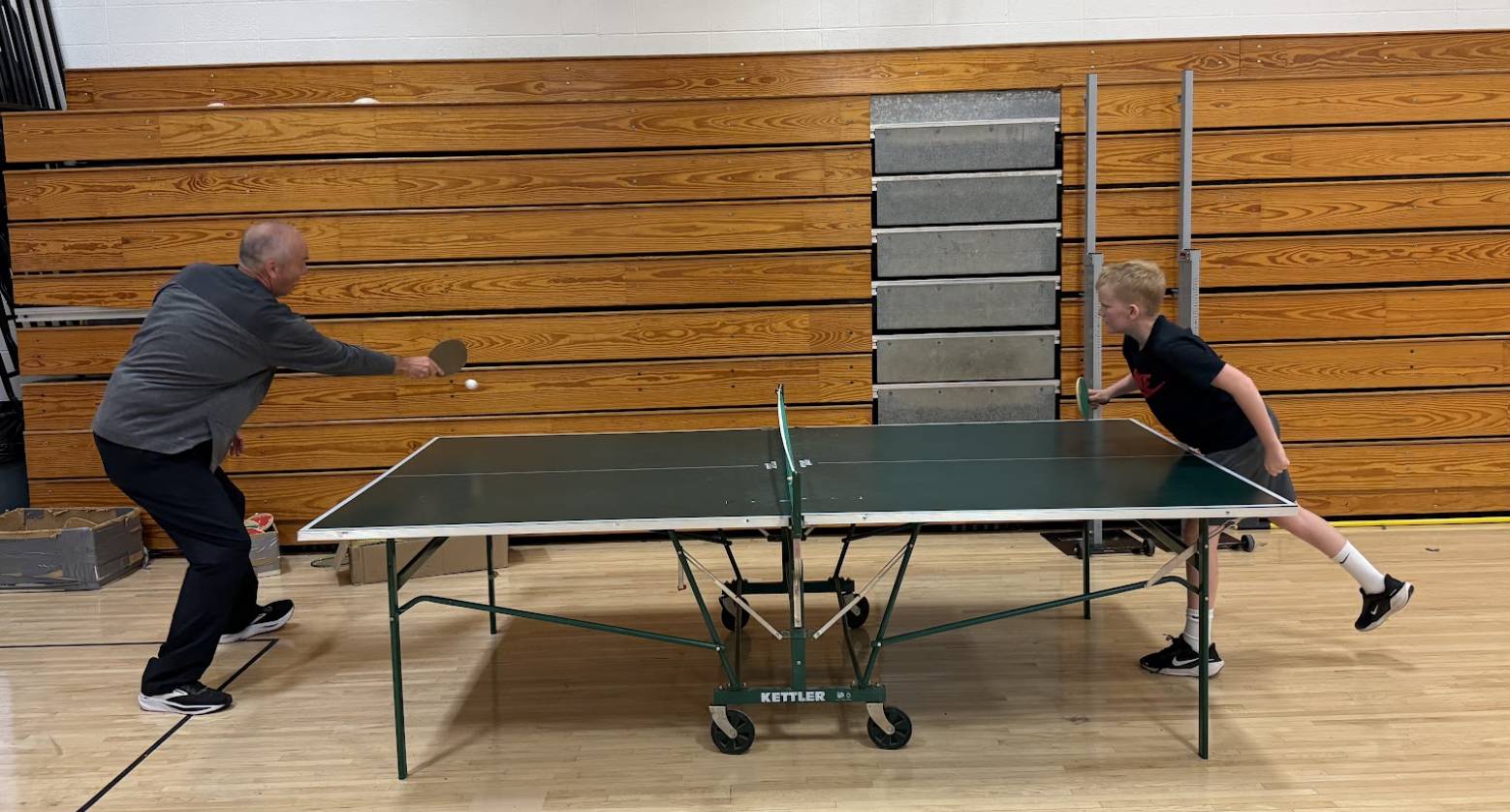 Adult and child play table tennis in a gym, leaning forward as they rally over a green ping-pong table.