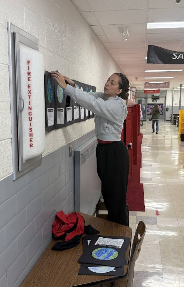 Woman stands on table in school hallway hanging student artwork