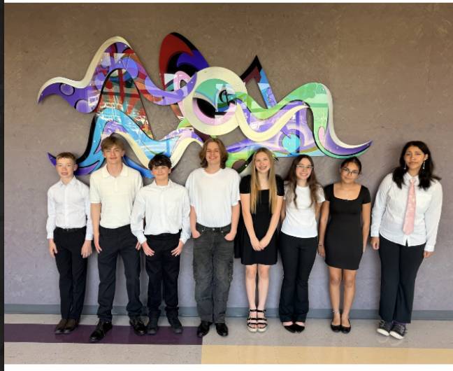 Nine students stand indoors in semi-formal attire beneath a colorful abstract wall sculpture, posing in a line.