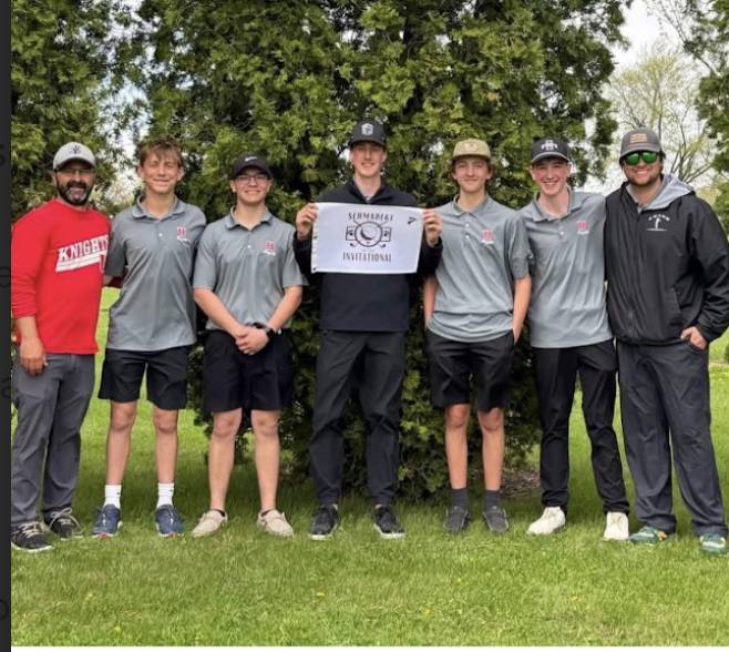 Seven teens and two adults stand on grass in front of trees; one holds a “Shamrock Invitational” sign, all smiling.