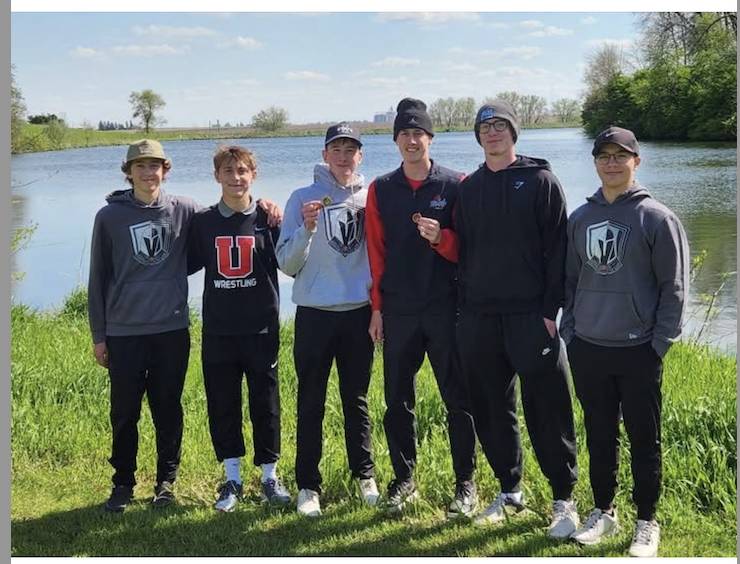 Five teenage boys stand beside a lake holding small metals during an outdoor fishing outing.