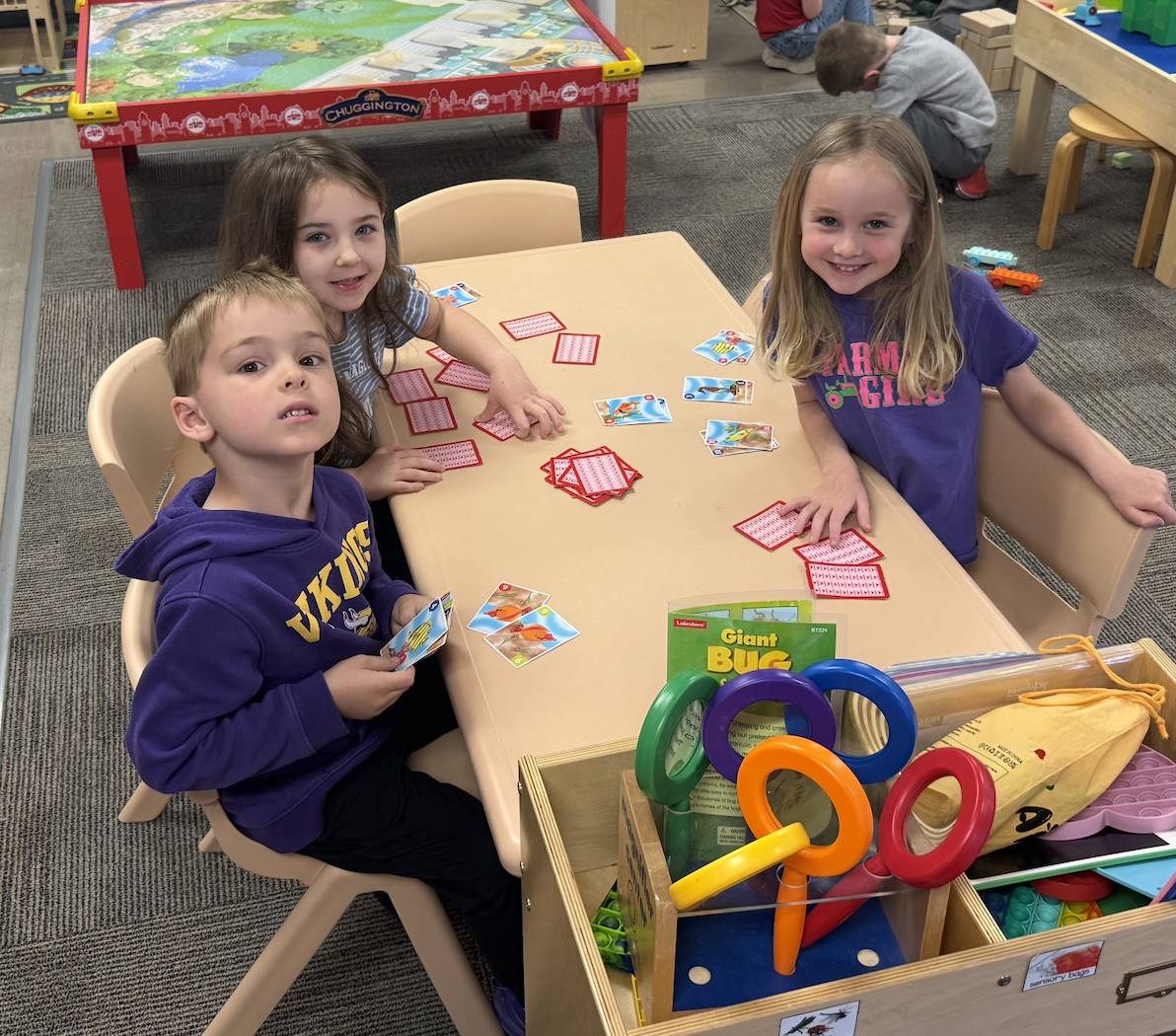 Three preschool students sit around a classroom table playing a matching card game with colorful learning tools nearby.