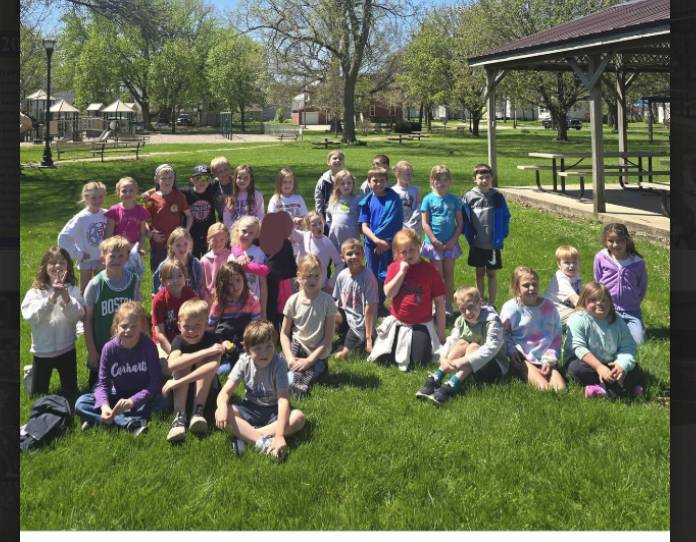 Elementary students pose together on a grassy park lawn near a pavilion and playground on a sunny day.
