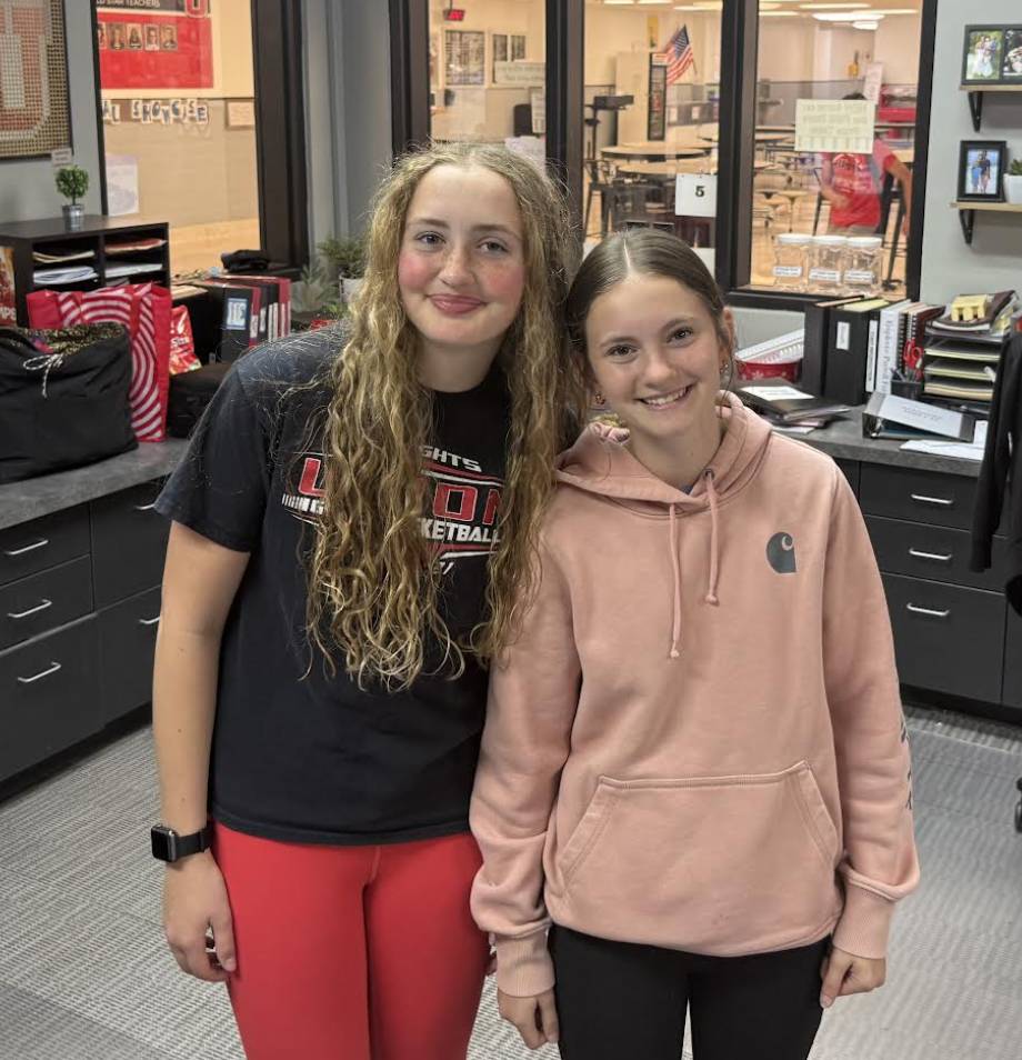 Two smiling students pose together inside a school office or classroom area.