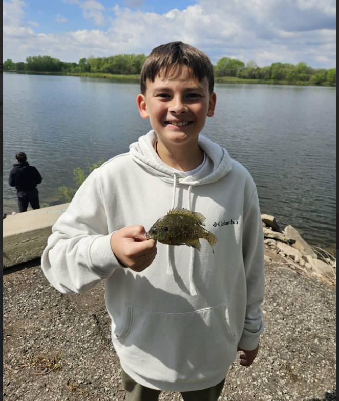 Student in a white hoodie smiles while holding a fish near a lakeshore with trees and water behind him.